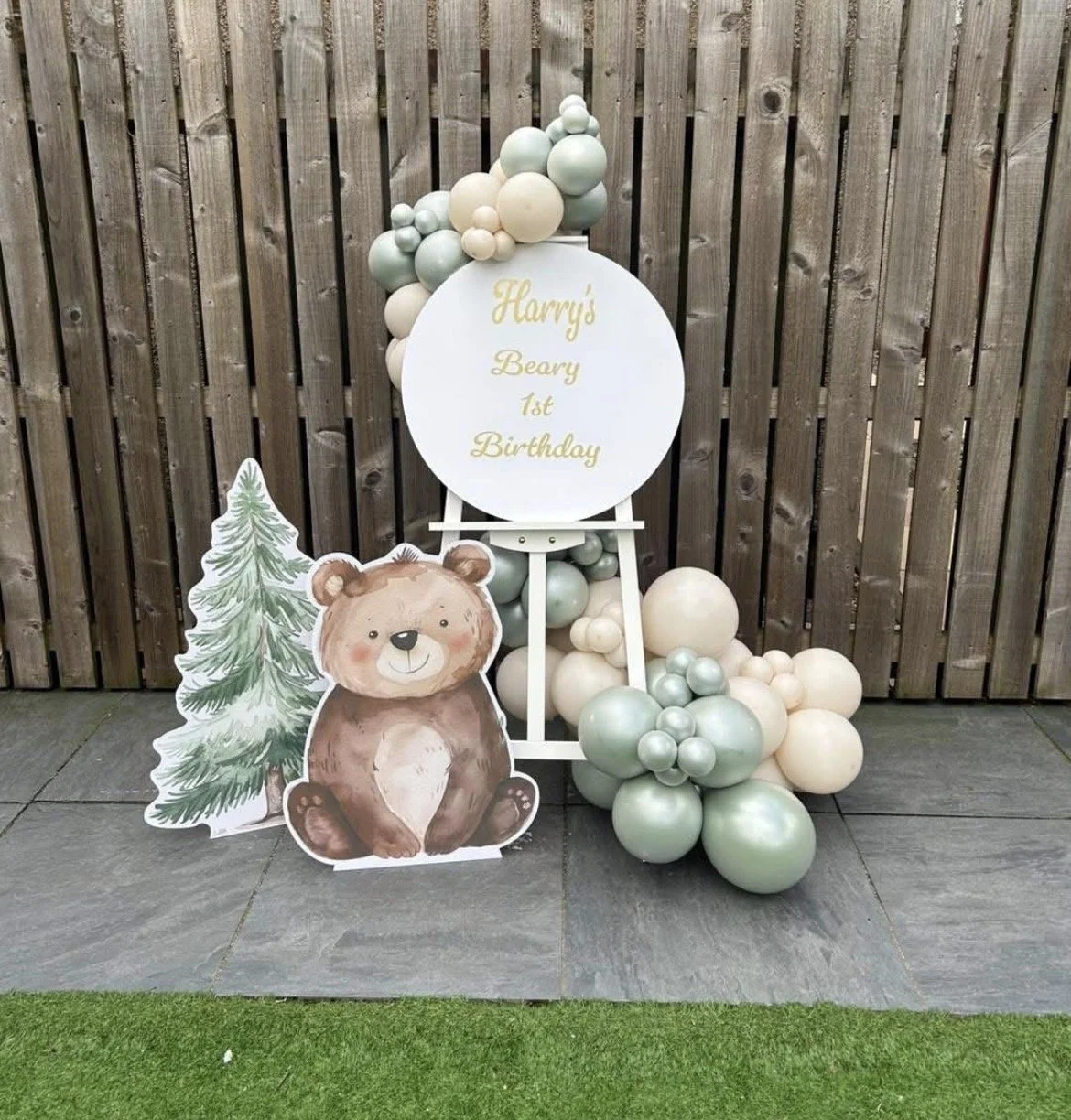 A birthday display with a wooden fenced background, decorated with pastel-colored balloons, a circular sign with gold text reading "Harry's Beary 1st Birthday," a paper cutout of a smiling teddy bear sitting next to a green Christmas tree illustratio