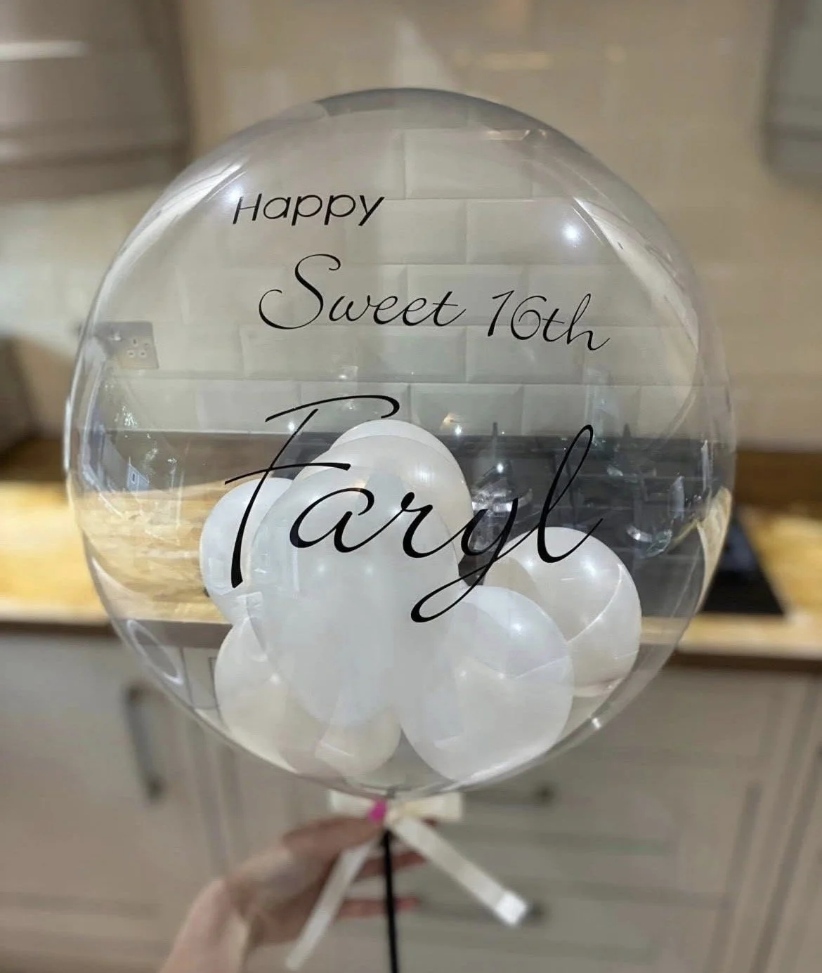 Clear balloon with white smaller balloons inside, decorated with black cursive text that reads 'Happy Sweet 16th Taryl', and a white ribbon at the bottom.