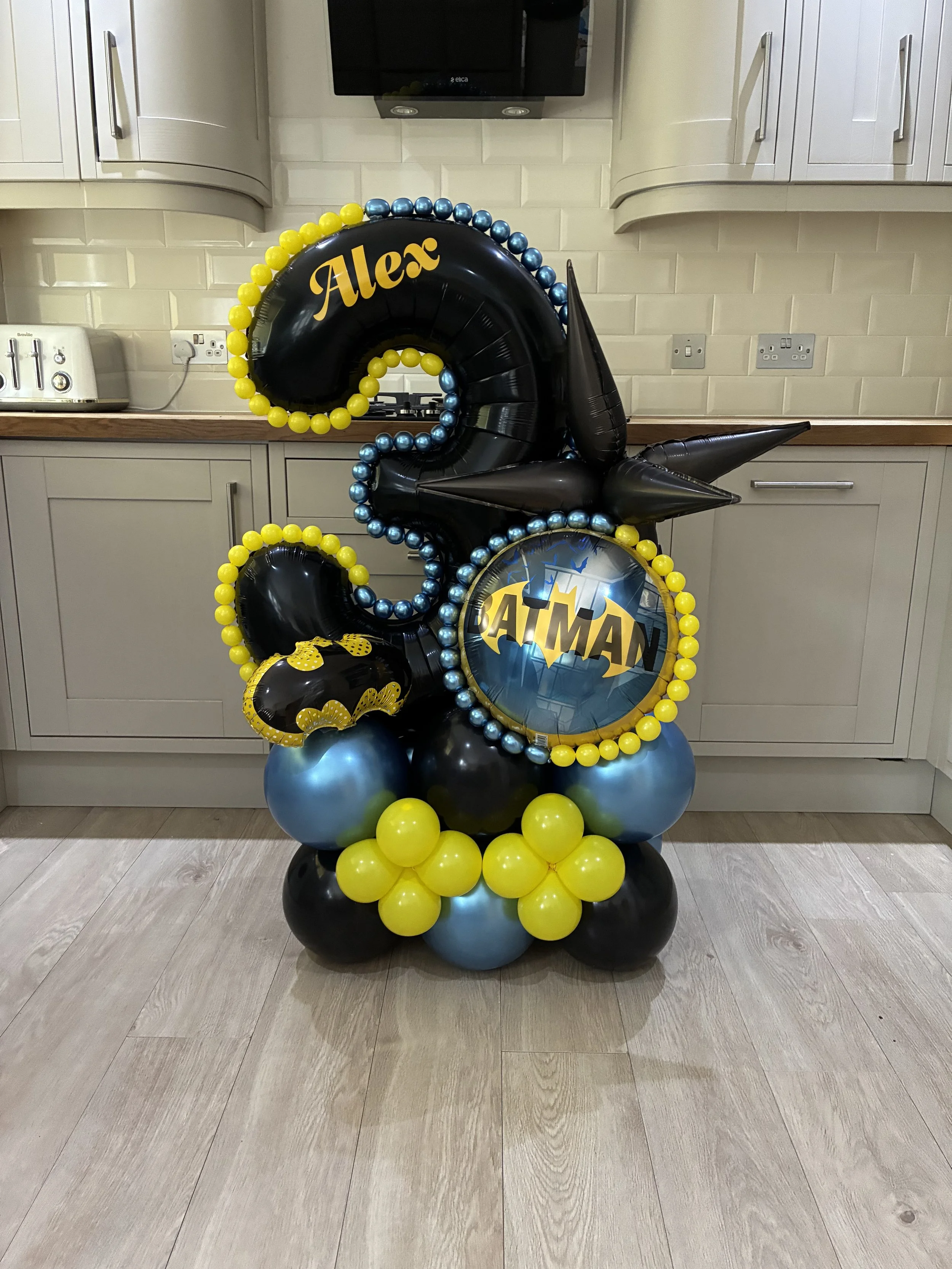 A birthday decoration display featuring black, yellow, blue, and Batman themed balloons, with the name 'Alex' on a black balloon, set in a kitchen with white cabinets and a wooden countertop.