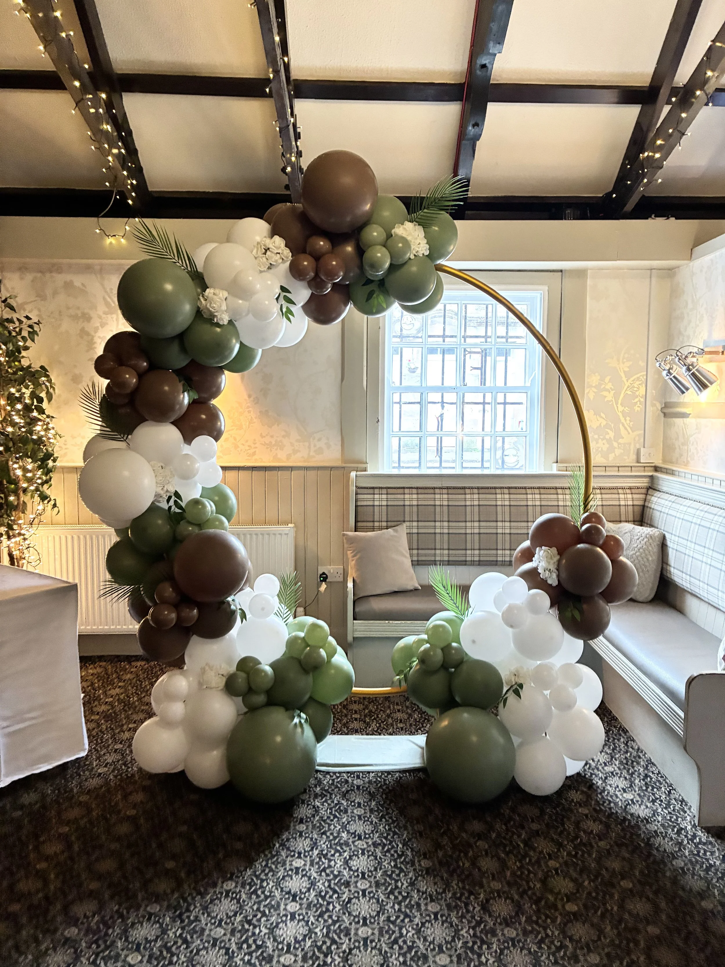 A balloon arch decorated with green, brown, and white balloons, with some balloons shaped like flowers and leaves, set up in a cozy indoor space with a sofa and window in the background.