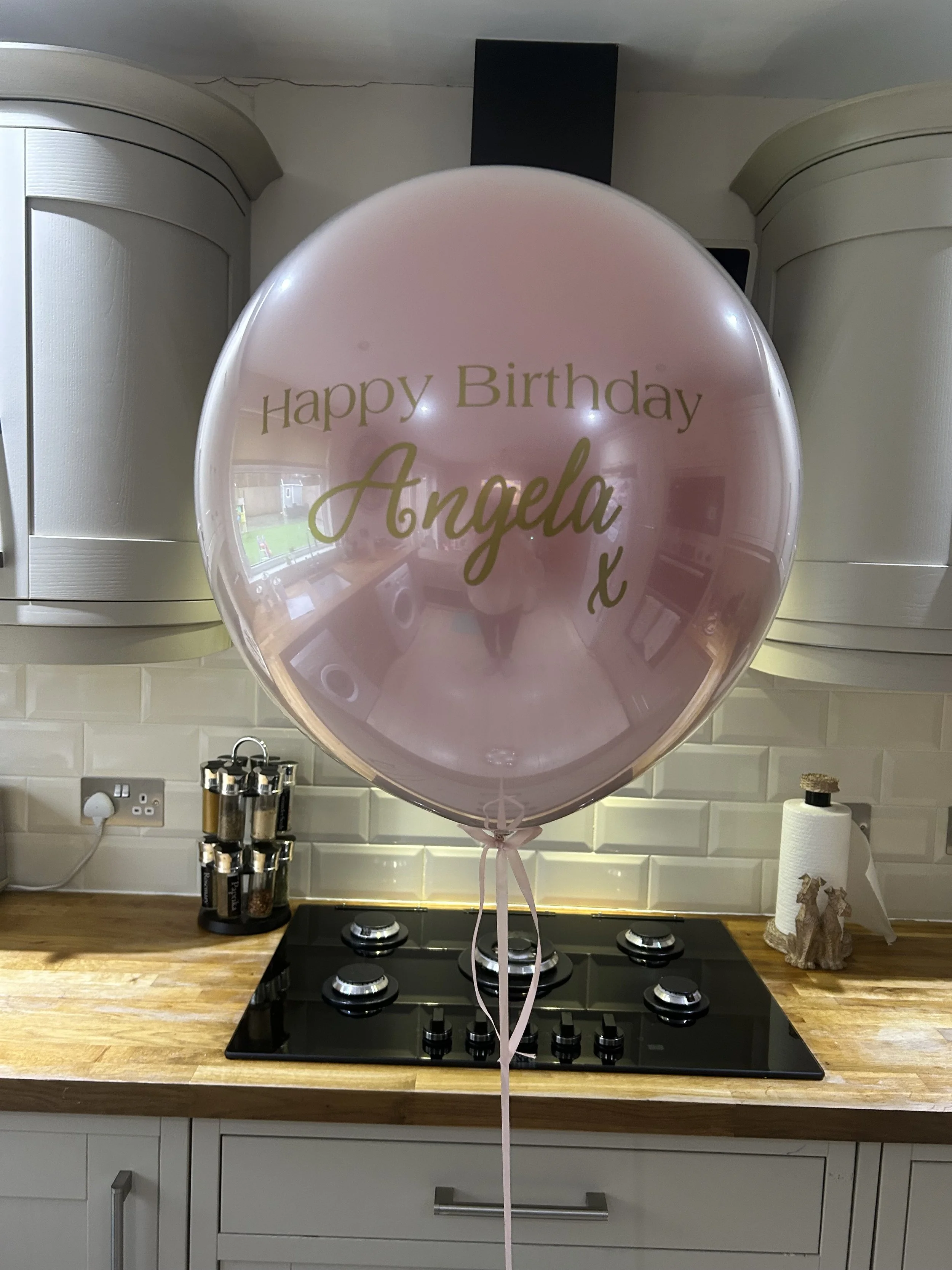 A pink balloon with gold text that reads 'Happy Birthday Angela' is tied with a pink ribbon and placed in front of a kitchen stovetop. The background features a white tiled backsplash, kitchen cabinets, a paper towel roll, and a utensil holder with s
