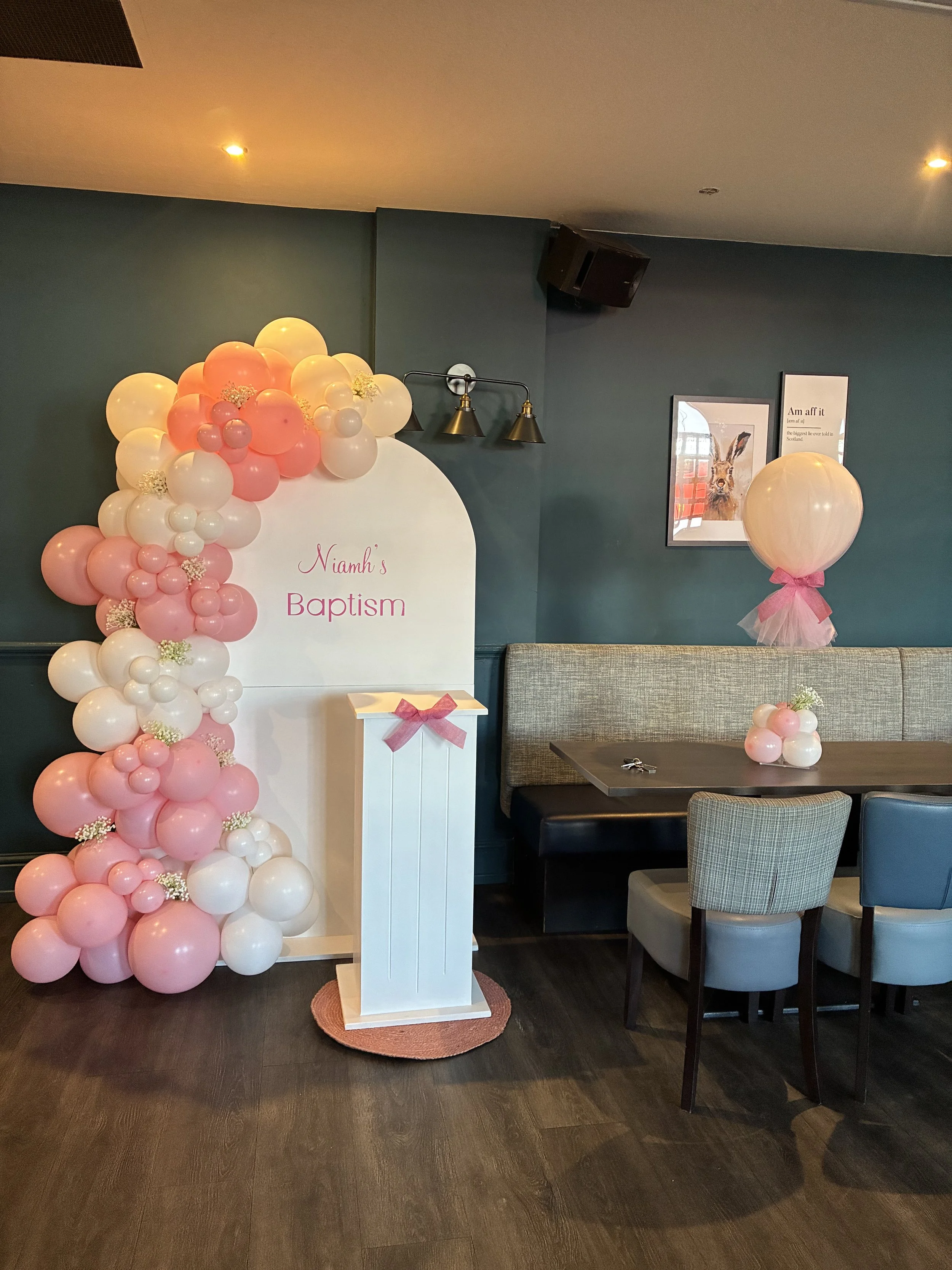 Inside a room decorated for a baptism with a white arch decorated with pink and white balloons and small flowers, a small white table with a pink bow, a gray bench, and a pink tulle balloon bouquet. There are framed pictures on the wall, including one of a rabbit, and the sign reads 'Niamh's Baptism.'