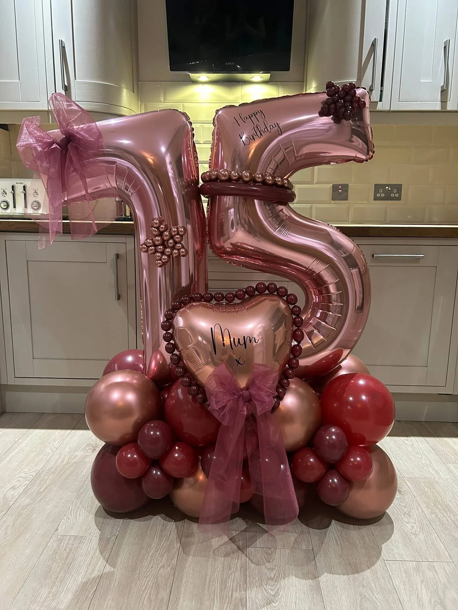 Decorative balloon display featuring large pink balloon shaped like a number '5' with the words 'Happy Birthday' written on it, surrounded by clusters of pink and red balloons, topped with a bunch of purple grapes, and pink ribbon, arranged on a wood