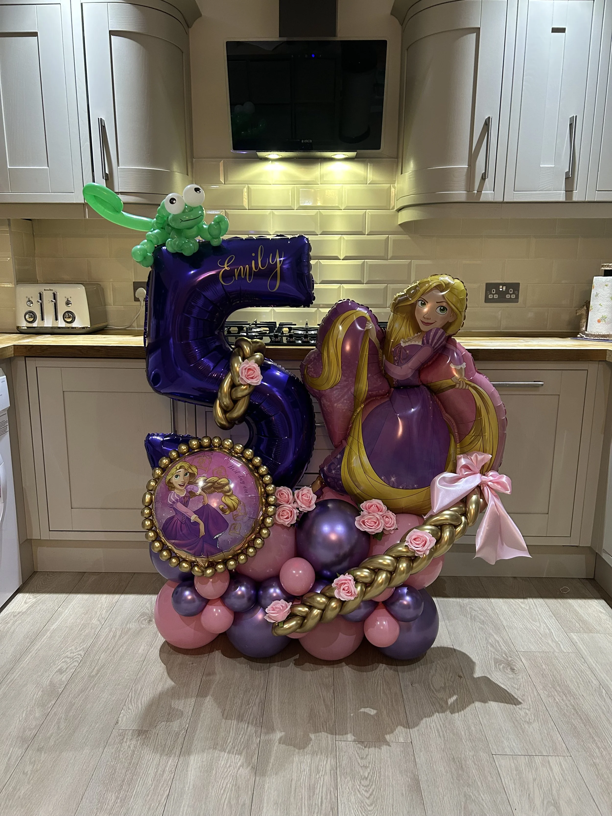 Decorative Rapunzel-themed birthday balloon arrangement with pink, purple, and gold balloons, including a large balloon of Rapunzel, a balloon of a purple number 5 with the name Emily, a green balloon animal, and pink roses, set in a modern kitchen with white cabinets and light-colored wooden flooring.