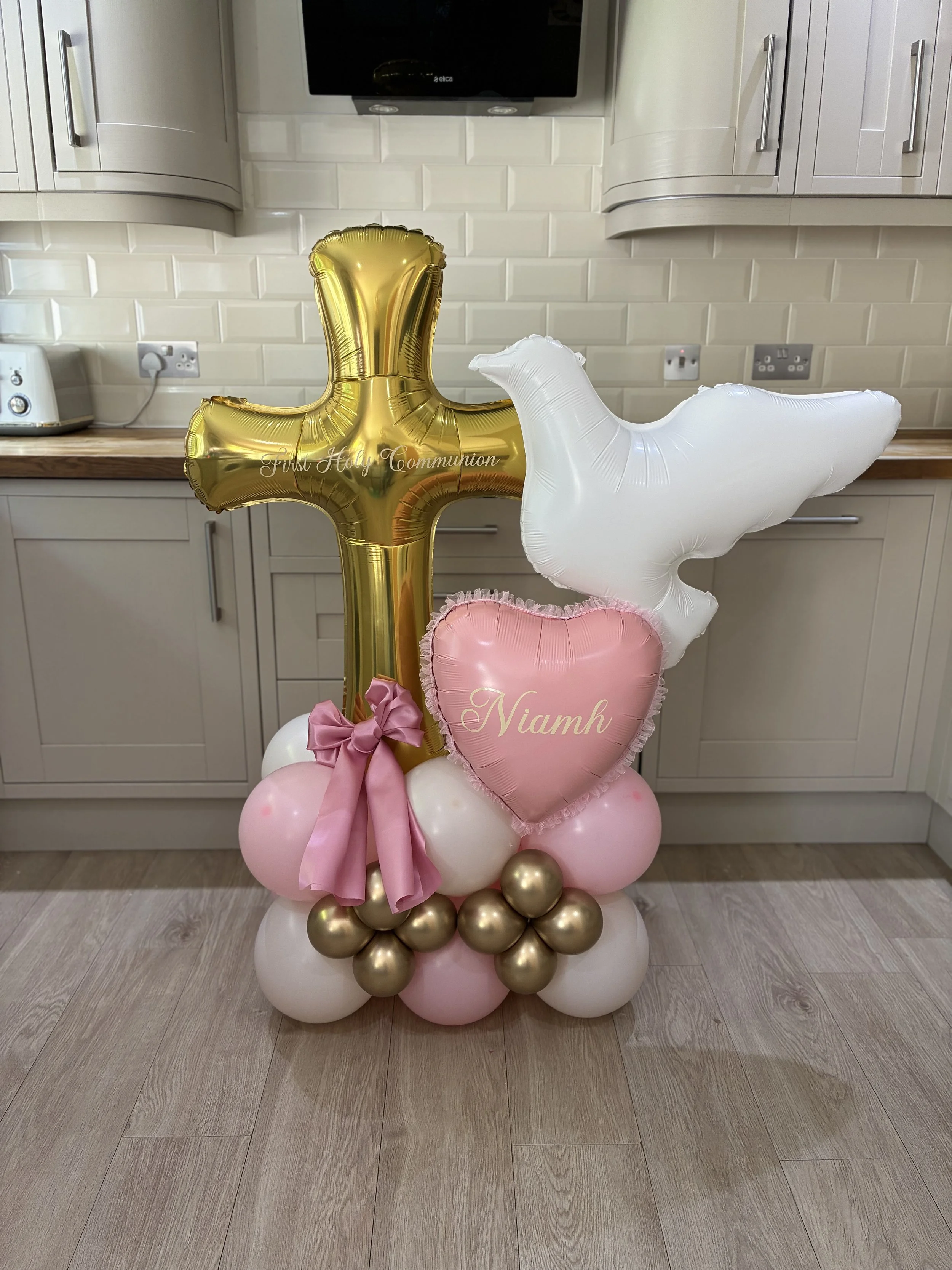 Balloon arrangement with gold and white balloons, pink heart-shaped balloon with the name 'Niamh', balloons arranged on the floor in a kitchen setting.