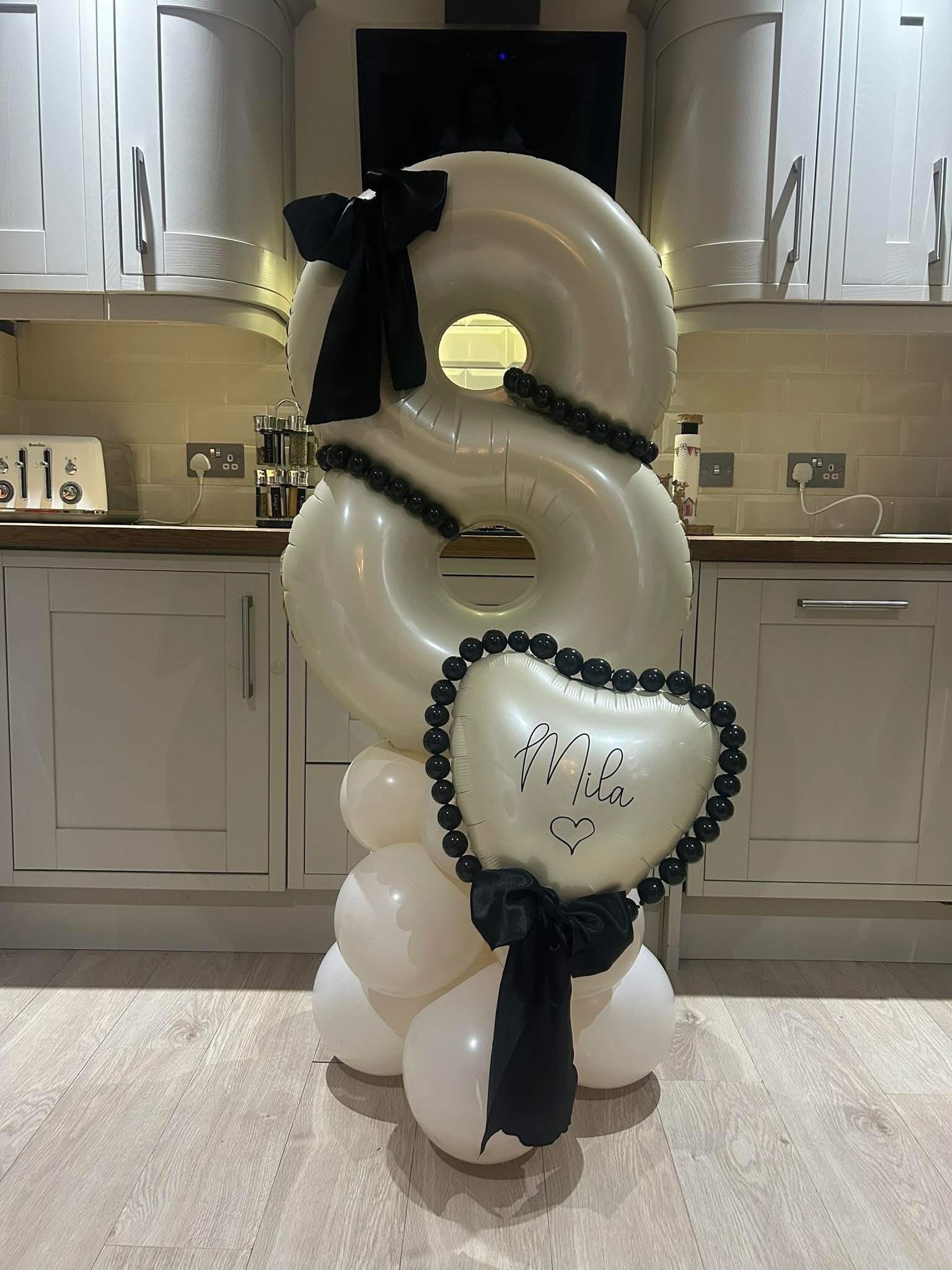 A decorative arrangement of white balloons with black accents, including a heart-shaped balloon inscribed with the name 'Mila,' placed in a home kitchen setting with white cabinets and beige brick backsplash.