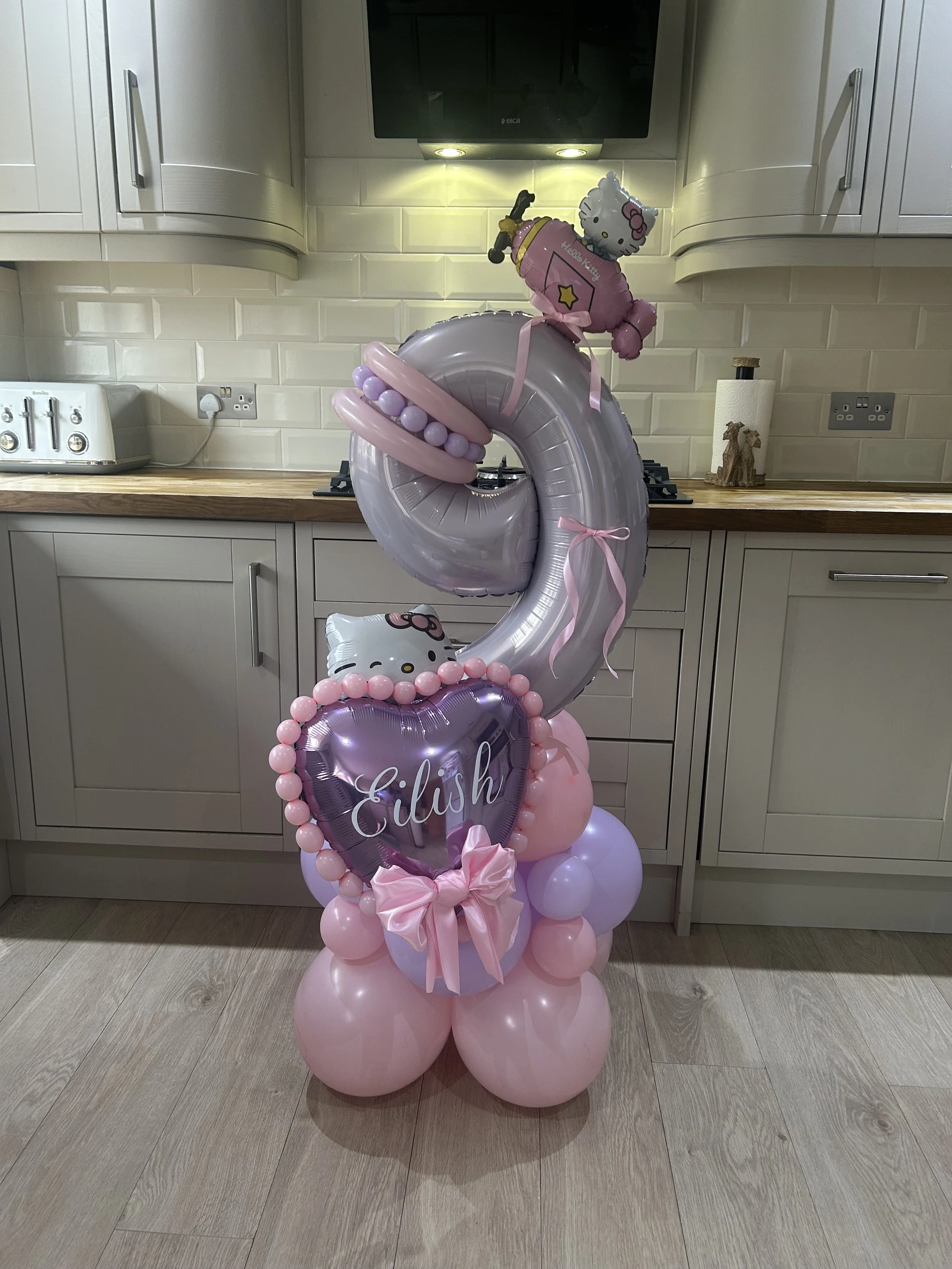 Pink and purple balloon arrangement with a Hello Kitty theme, including a large number '2' balloon, a balloon with Hello Kitty's face, and a balloon with 'Eilish' written on it, decorated with pink ribbons and smaller pink and purple balloons.