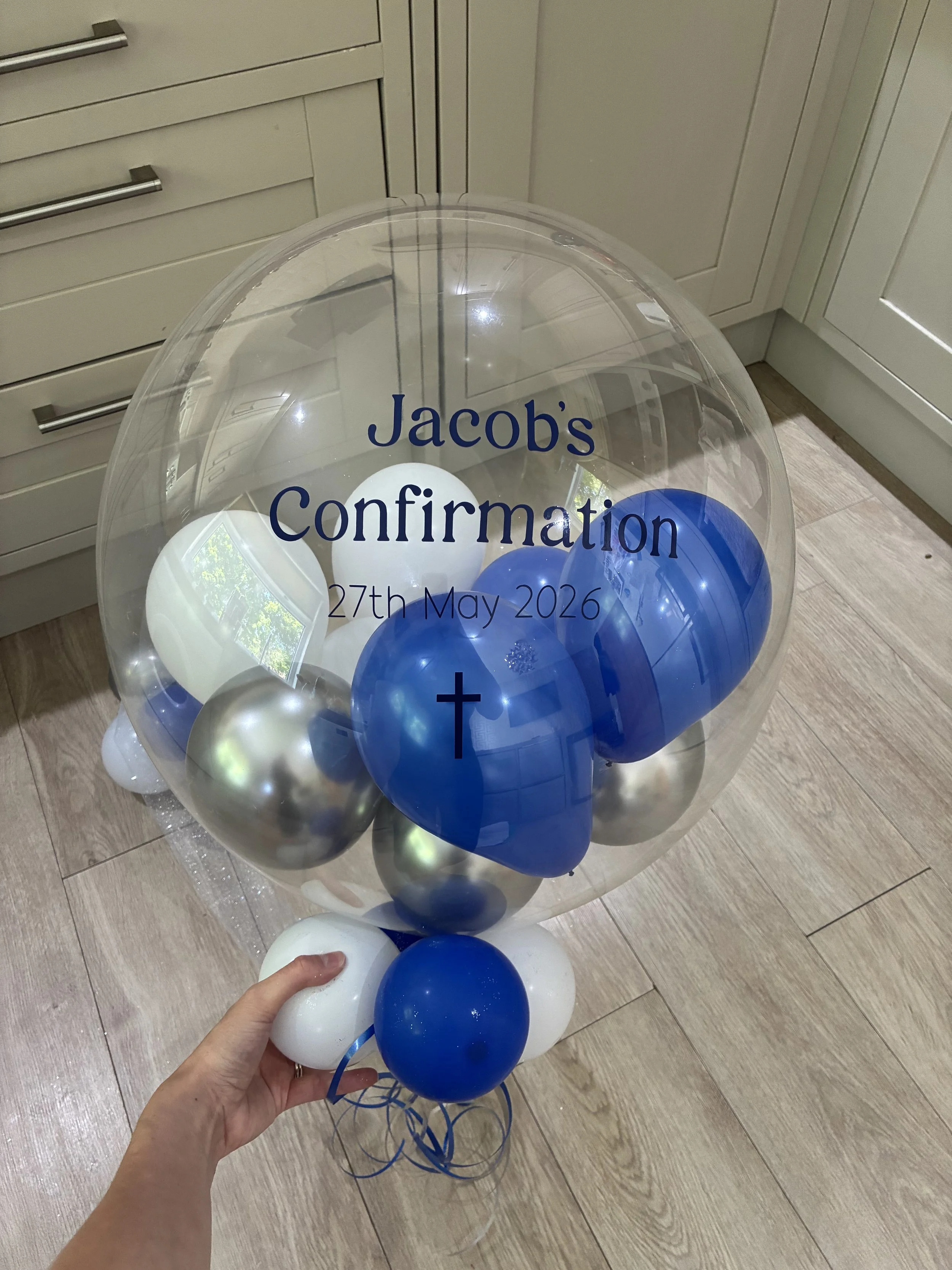 A clear balloon filled with blue, white, and silver balloons, with text that reads 'Jacob's Confirmation, 27th May 2026', being held by a person in a room with wooden flooring and cream-colored cabinets.