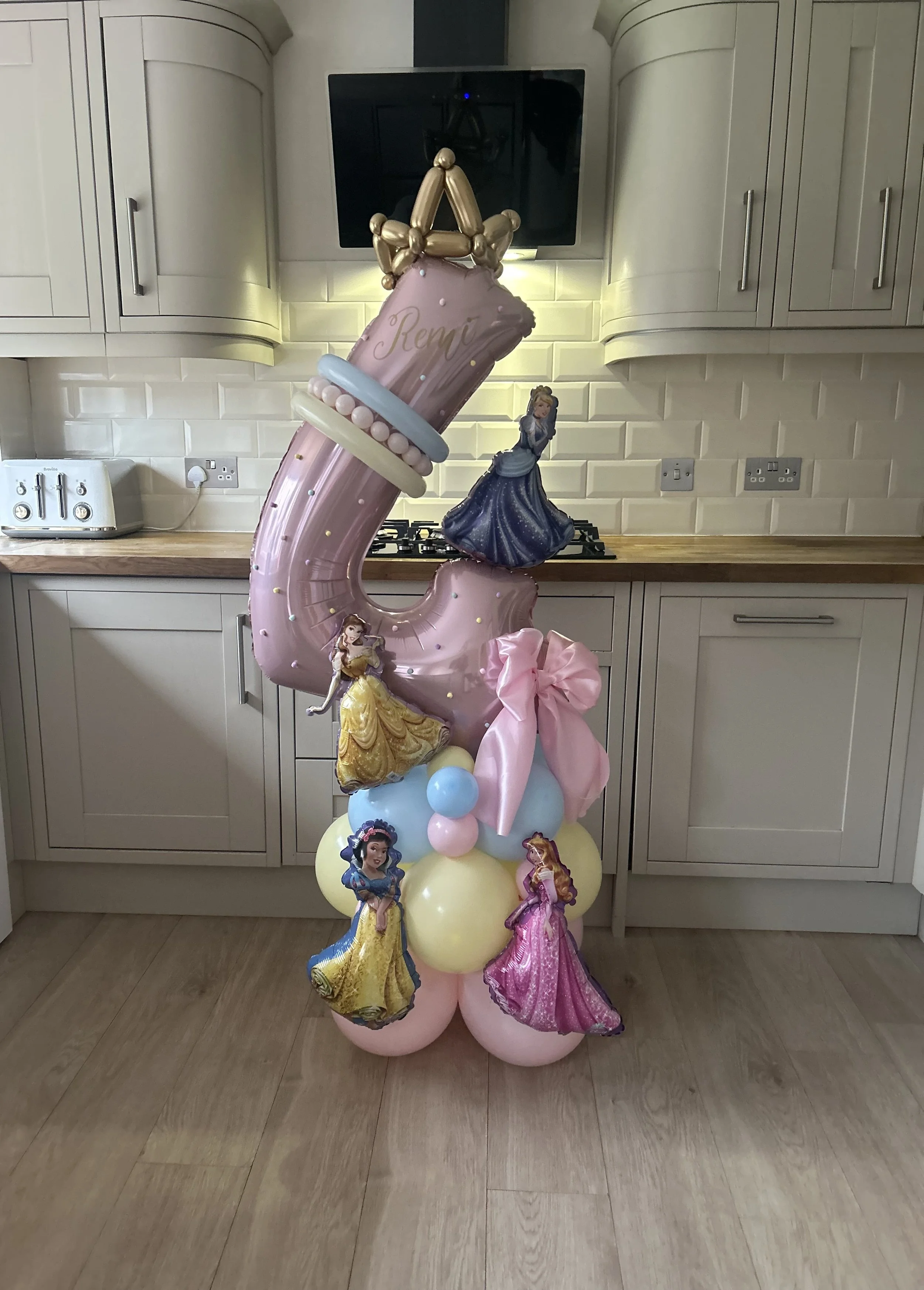 Balloon display with Disney princess themes, including Belle, Cinderella, Aurora, and Snow White, with a large pink balloon number '1' at the top, situated in a kitchen with white cabinets and wooden countertops.