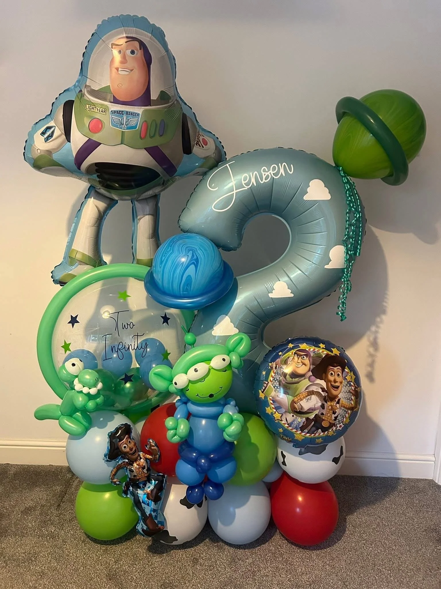 A colorful balloon display featuring characters from Toy Story, including Buzz Lightyear, Woody, and aliens, with balloons spelling 'Jensen' and 'Two Infinity' and decorated with planets, stars, and space-themed elements.