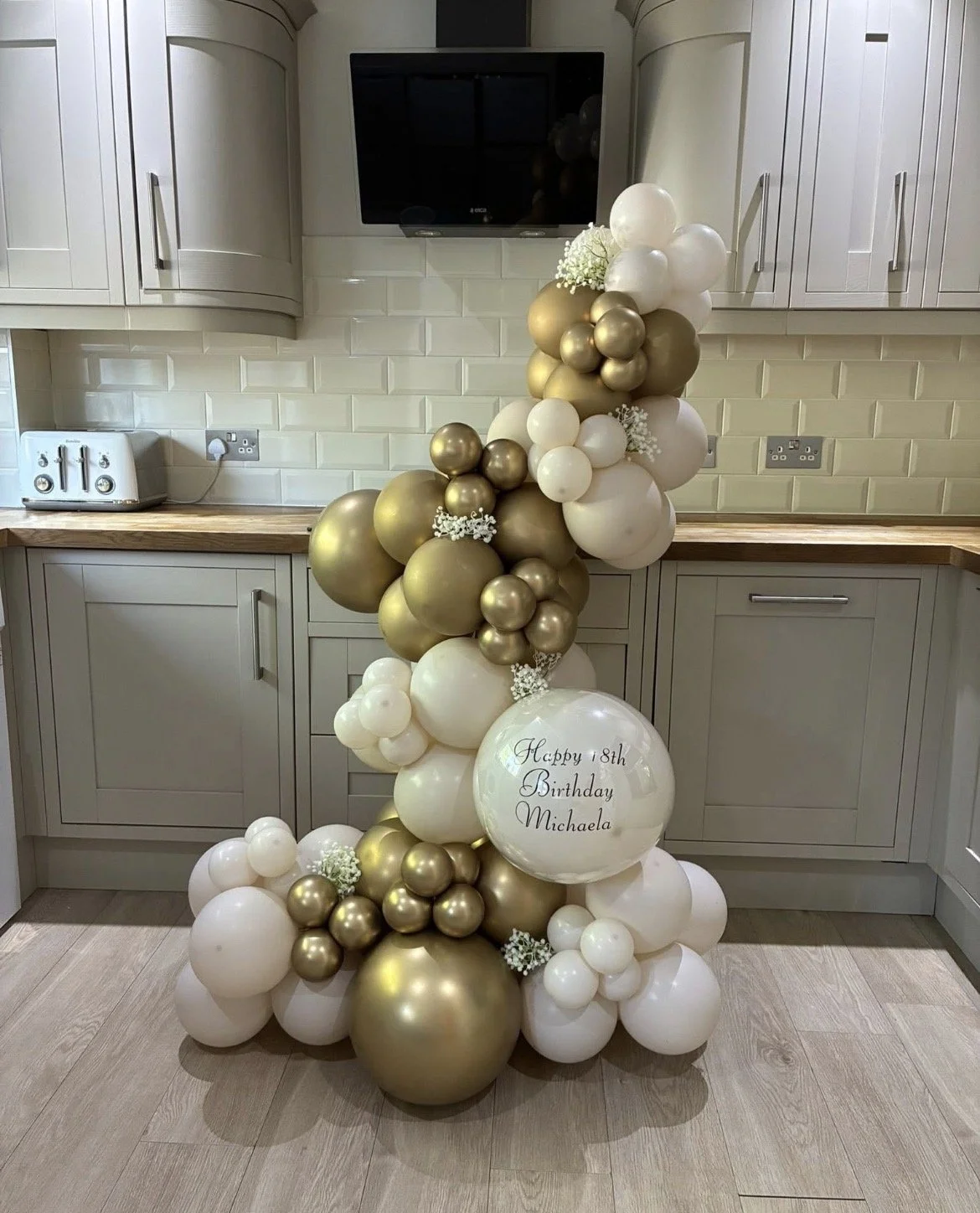 A balloon arch in gold, white, and cream colors with small white flowers, featuring a large sphere with a birthday message for Michaela's 18th birthday in a kitchen.