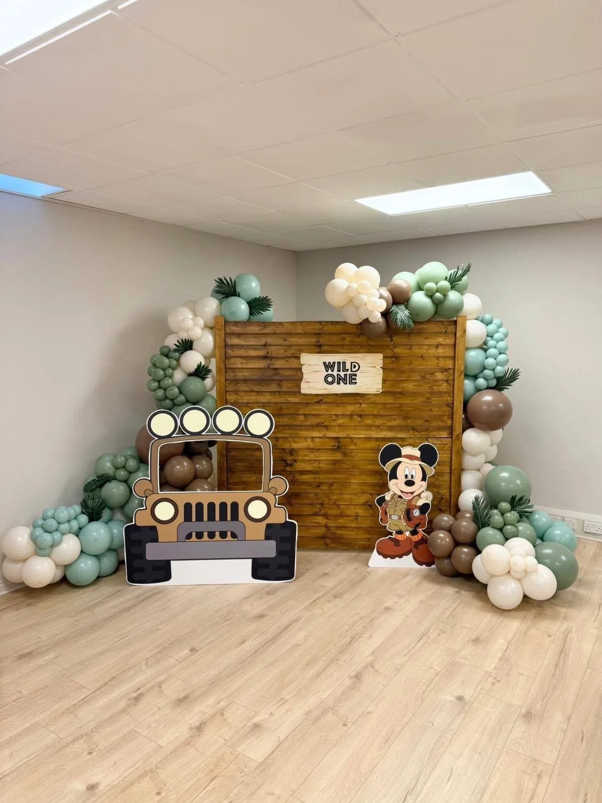 Decorative jungle party setup with balloon arch in green, white, and brown, featuring a wooden backdrop with a "Wild One" sign, a cardboard cutout of a jeep, and a cartoon Mickey Mouse dressed as an explorer.