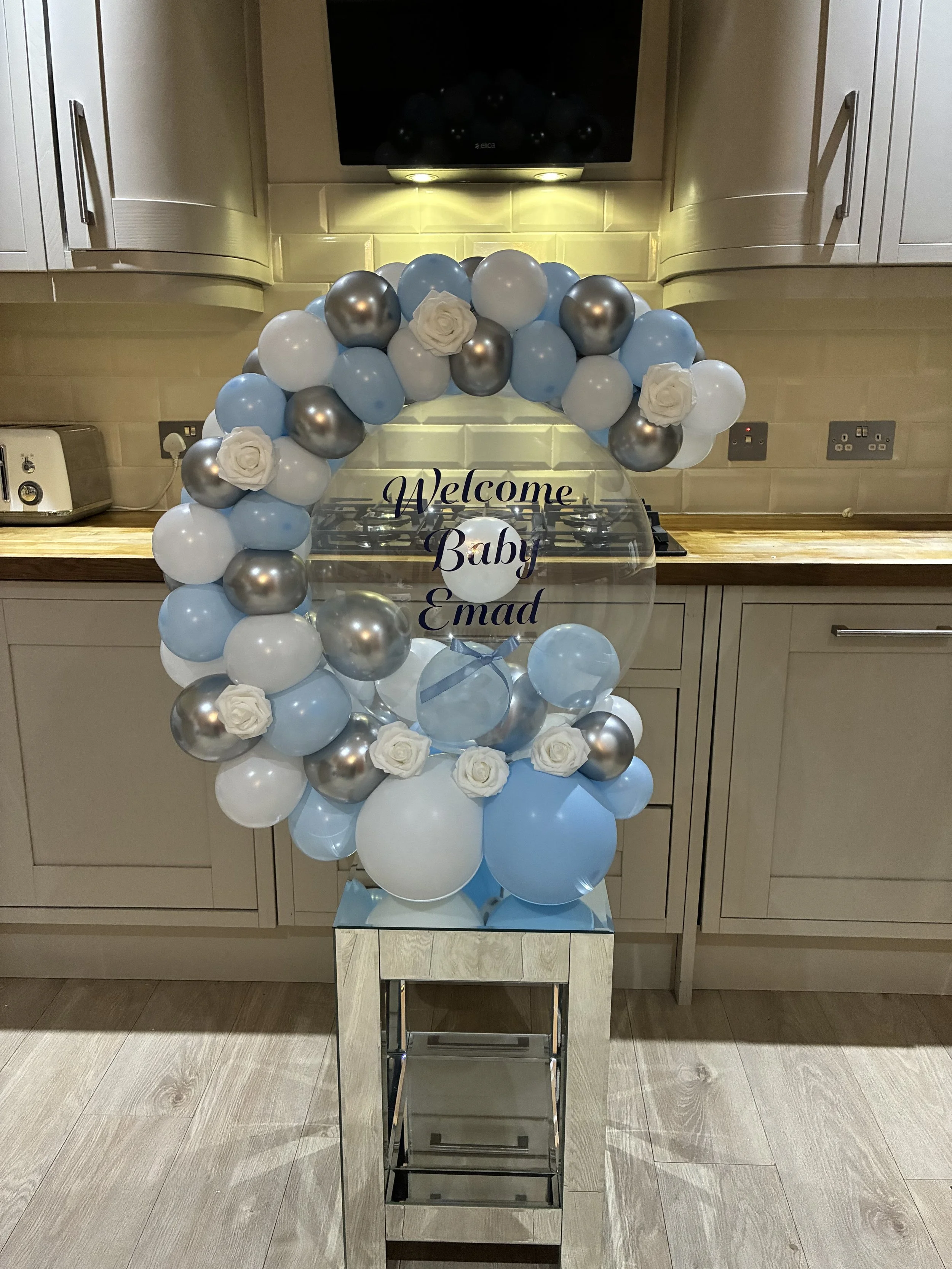 A balloon arch with white, blue, and metallic silver balloons, decorated with white roses, surrounds a glass sign that reads 'Welcome Baby Emad' in a kitchen. The arch is on a reflective stand on the floor.