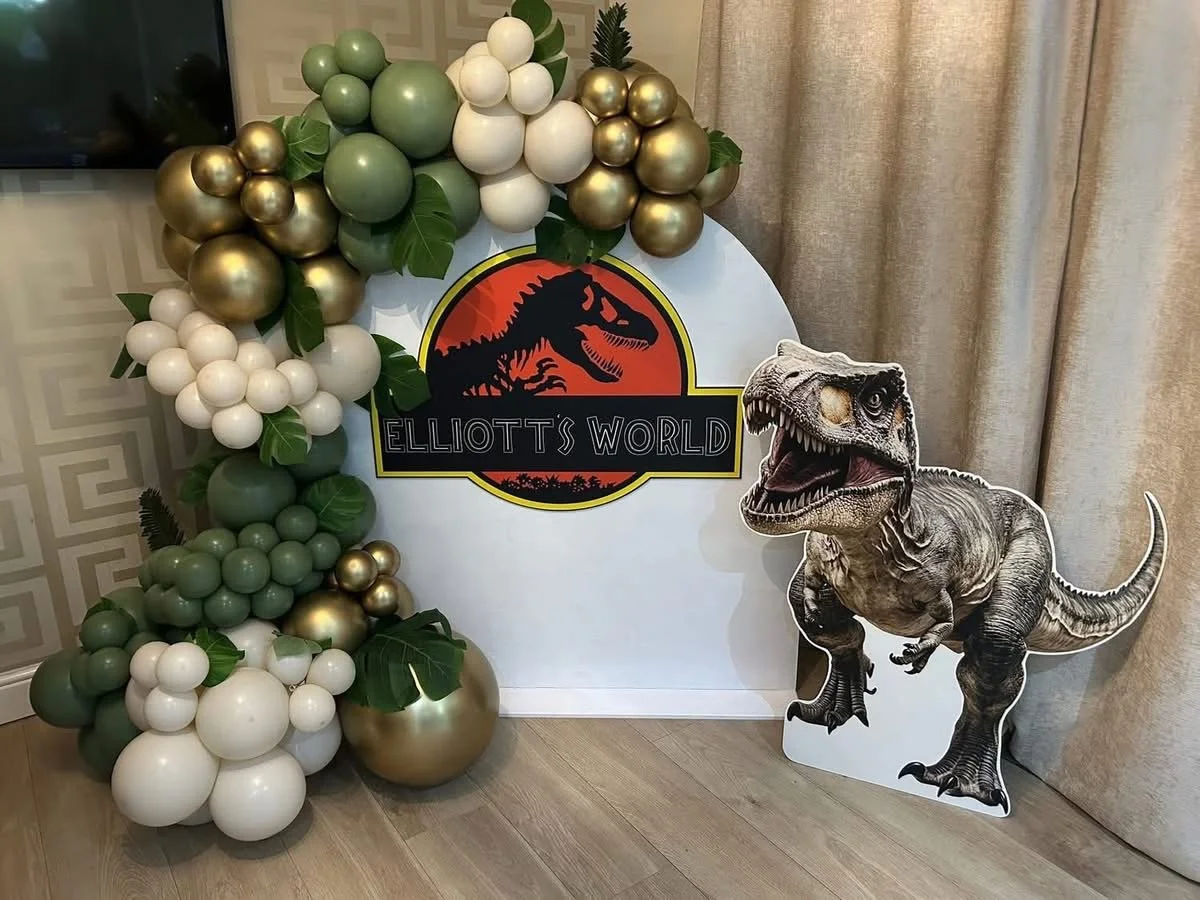 Decorative display with balloons and a Jurassic Park themed sign reading "Elliott's World". Includes a large dinosaur cutout and clusters of green, white, and gold balloons with leaves, next to a curtain and wall with a geometric pattern.