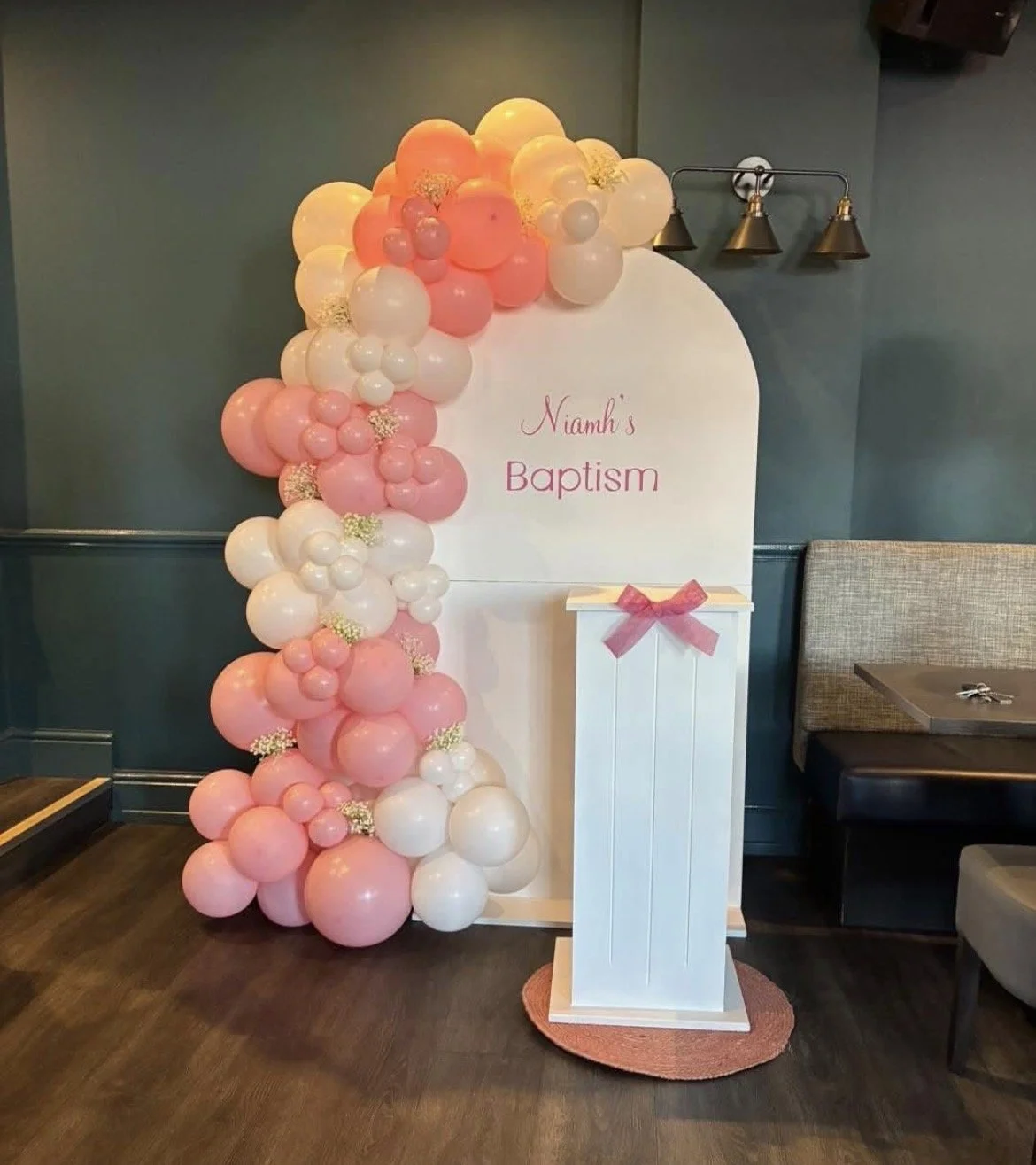 Decorative setup for a baby's baptism with a white sign reading 'Niamh's Baptism' surrounded by pink and white balloons on the left side and a small white stand with pink bow on the right.