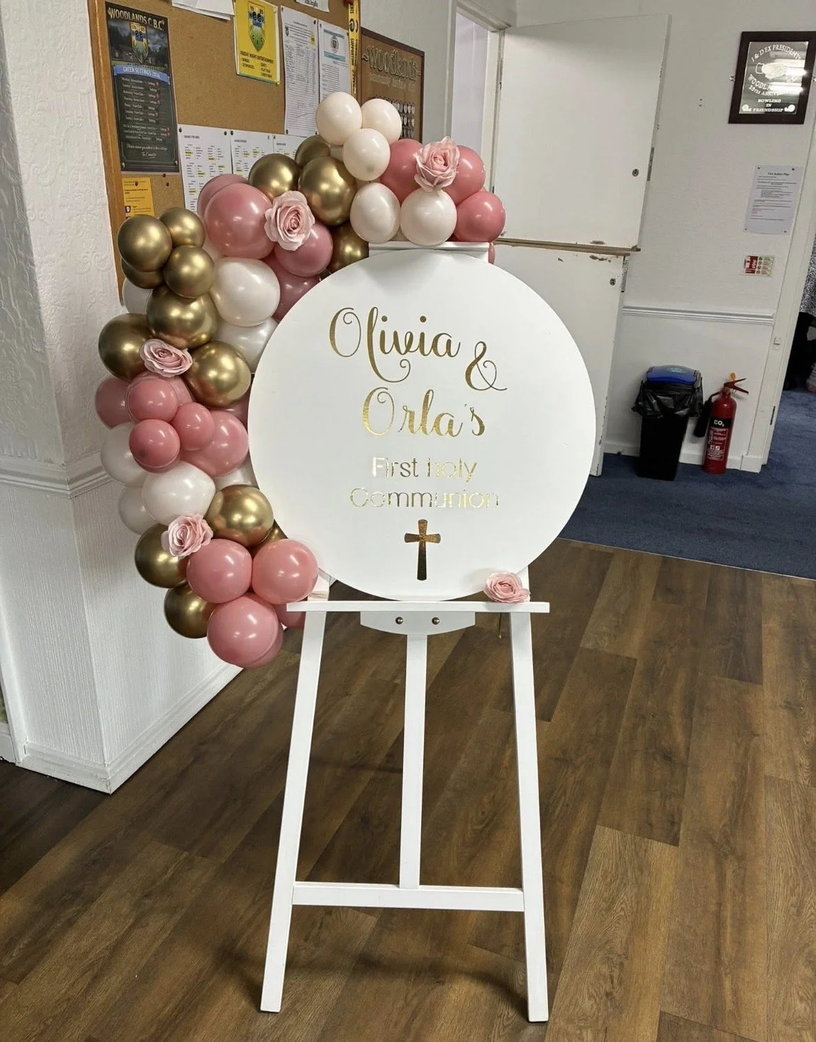 Celebration display for Olivia and Orla's First Holy Communion, featuring a white round sign with gold lettering on an easel, surrounded by pink, white, and gold balloons, and decorated with pink artificial roses.