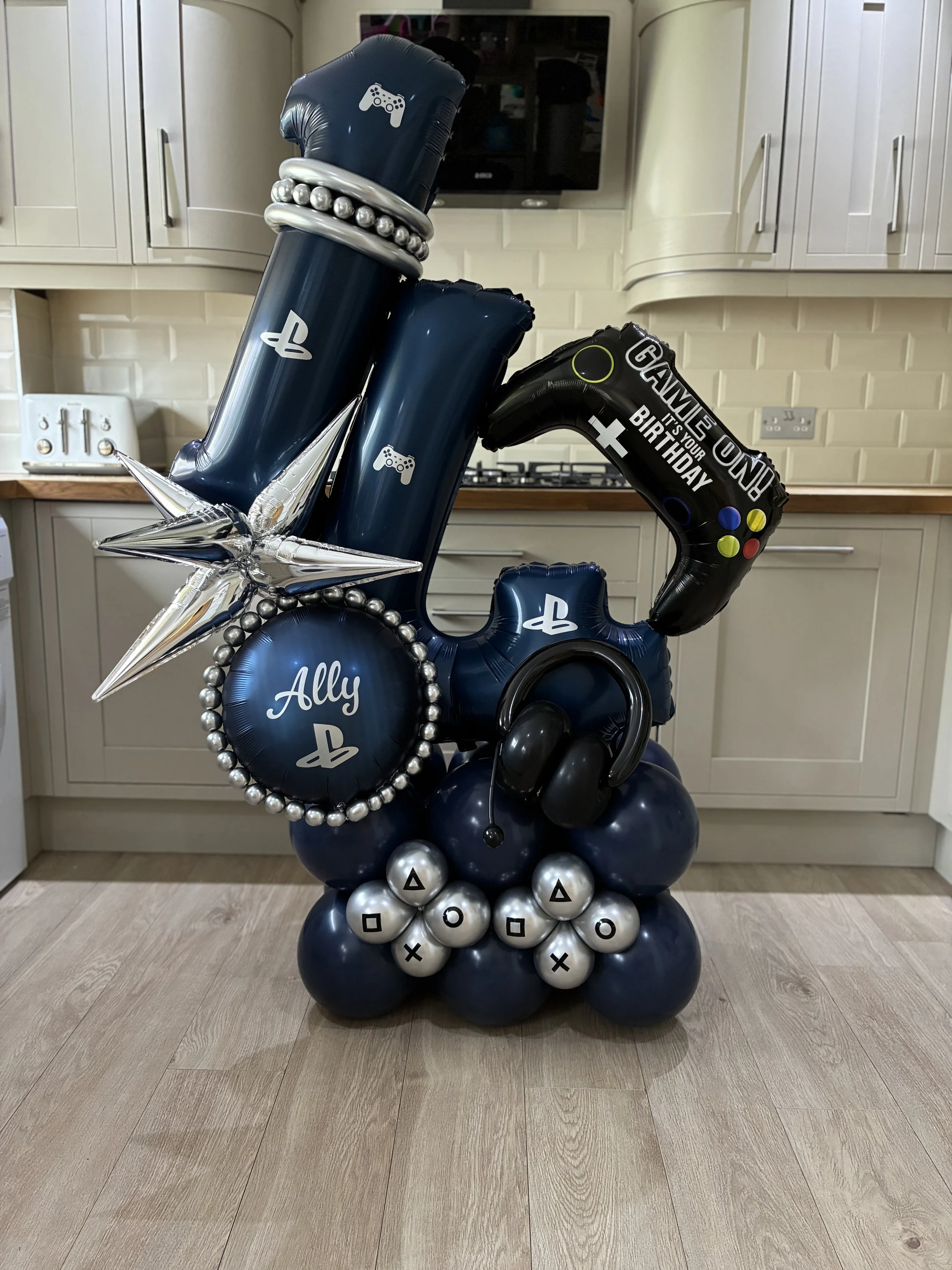 A balloon sculpture featuring PlayStation controller themed balloons, a large silver star, and a sign with birthday message, arranged on a base of black and silver balloons in a room with kitchen cabinets in the background.