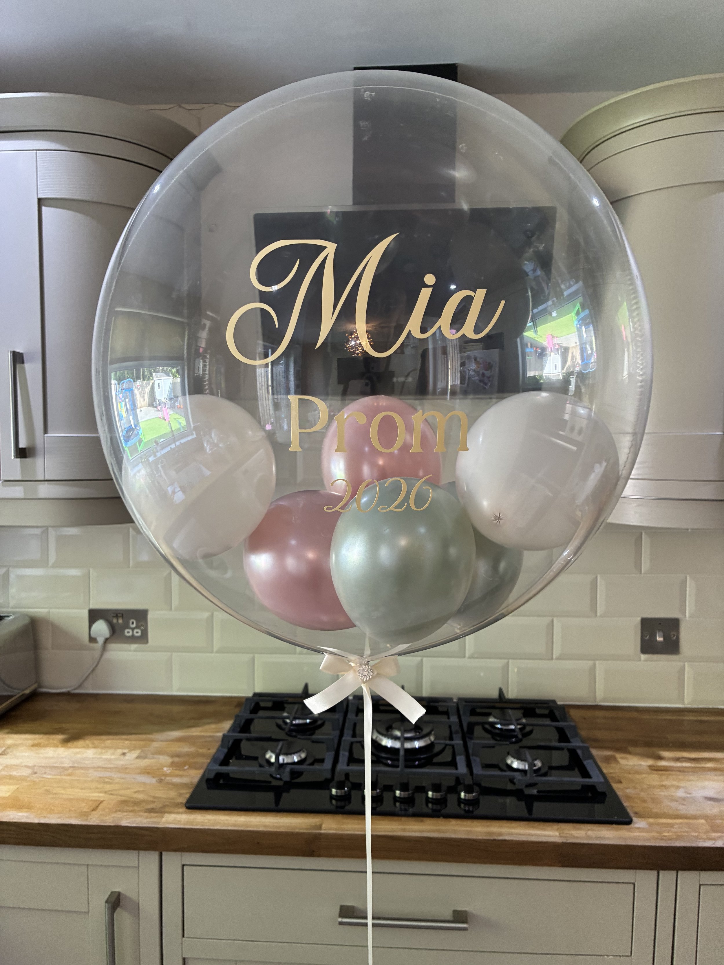A large transparent balloon with the words 'Mia Prom 2026' in gold lettering, filled with smaller balloons in shades of pink, gold, and white, tied with a white ribbon at the bottom, sitting on a kitchen countertop in front of a stove.