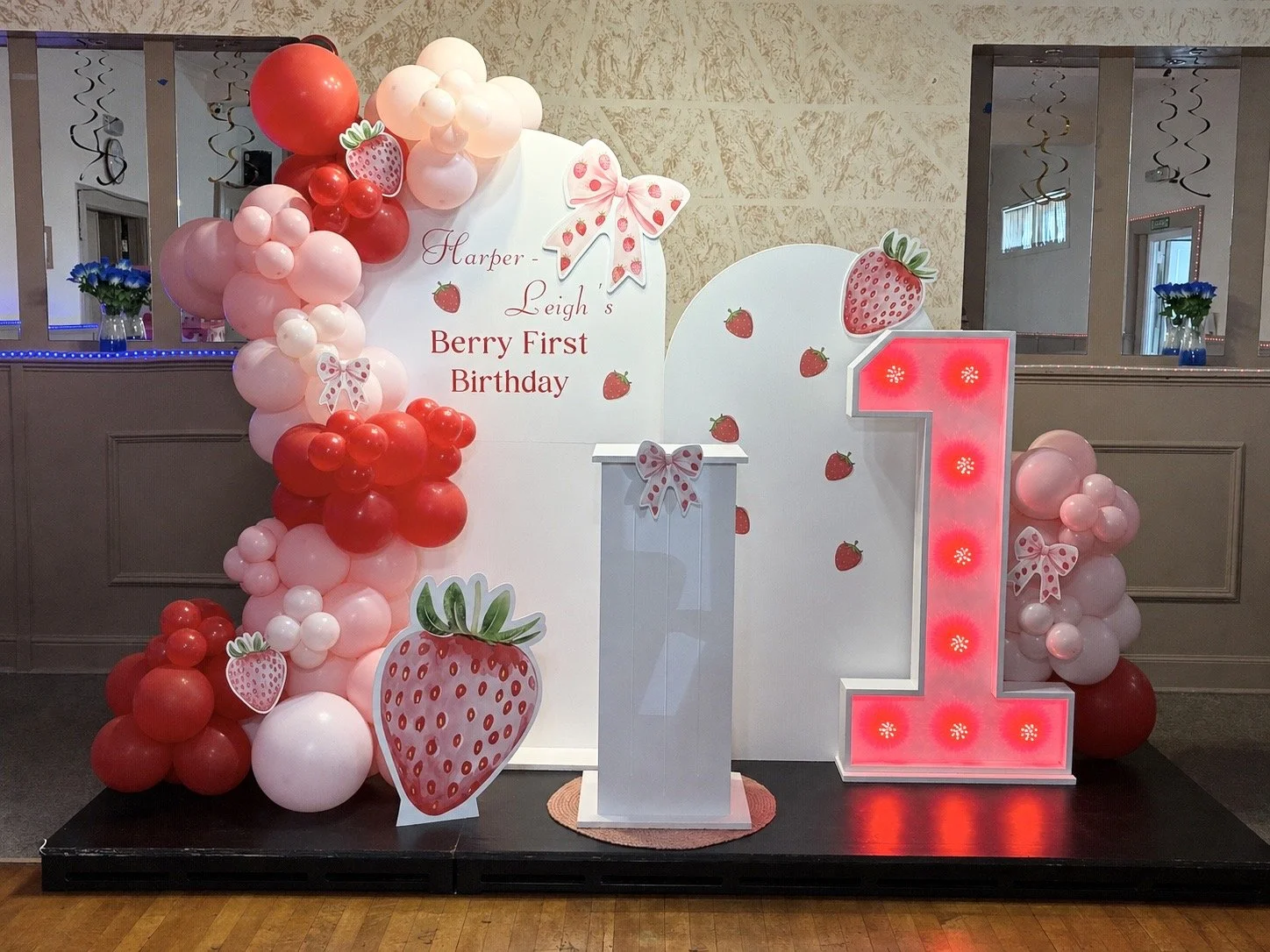 Decorations for a first birthday party with strawberry theme, featuring balloons, a large illuminated number one, and a backdrop with strawberries and text.