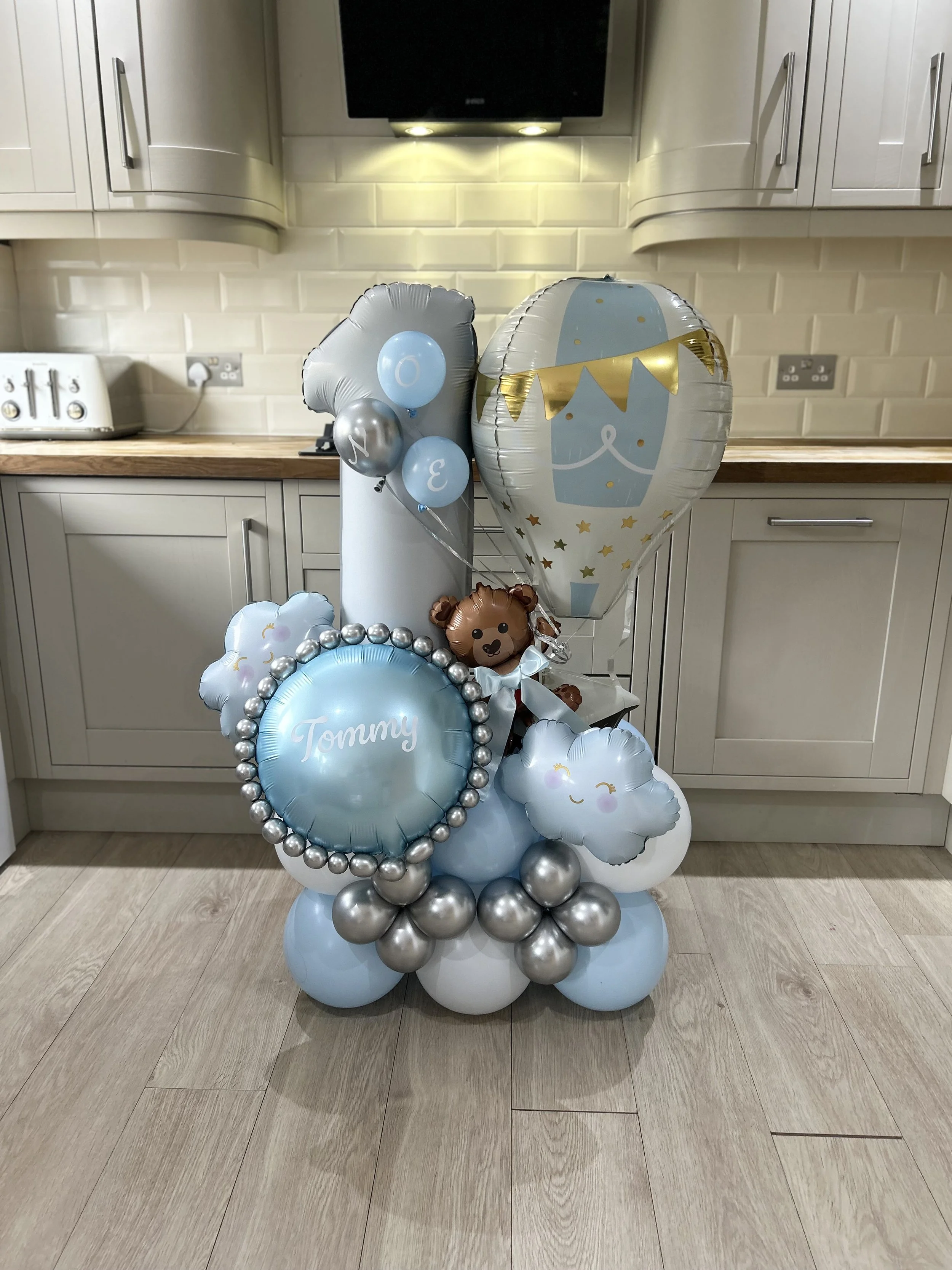 A cake shaped balloon arrangement with balloons that form a baby shower theme, including a large balloon with a baby face and a bunting, one with the name 'Tommy', and other balloons with clouds and stars, set in a kitchen with cream-colored cabinets