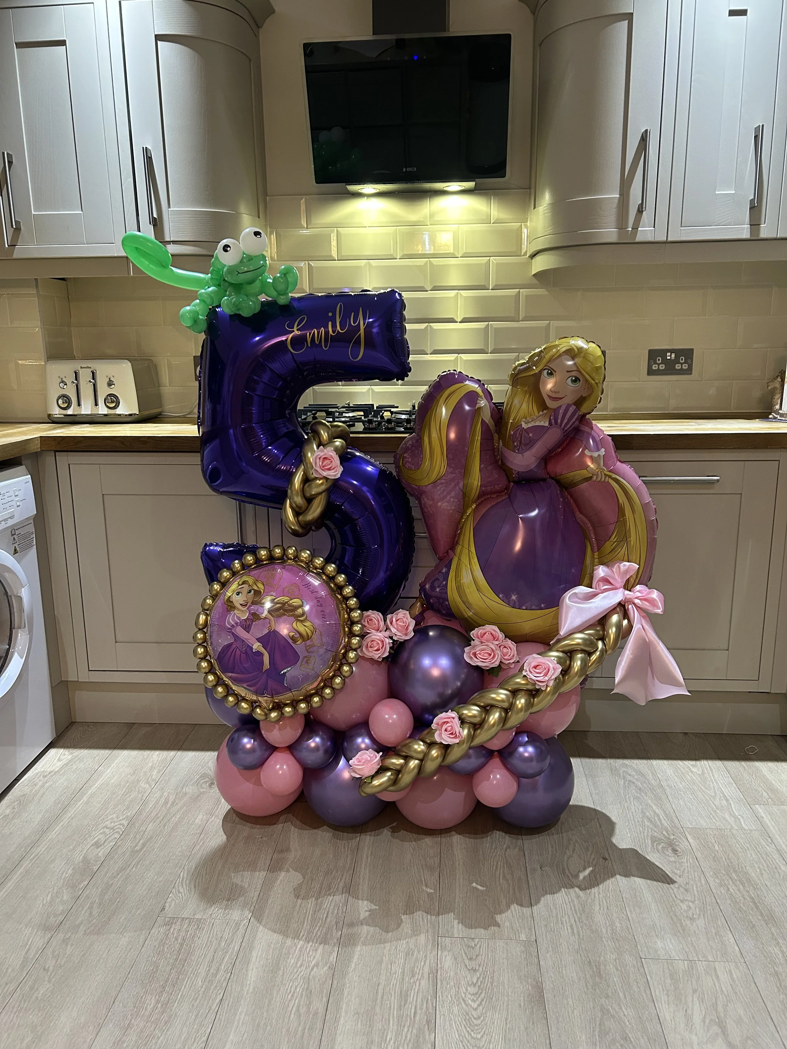 Balloon arrangement featuring Rapunzel from Disney's Tangled, pink roses, purple, pink, and gold balloons, including a large purple number 5 balloon with the name Emily written on it, set in a kitchen.