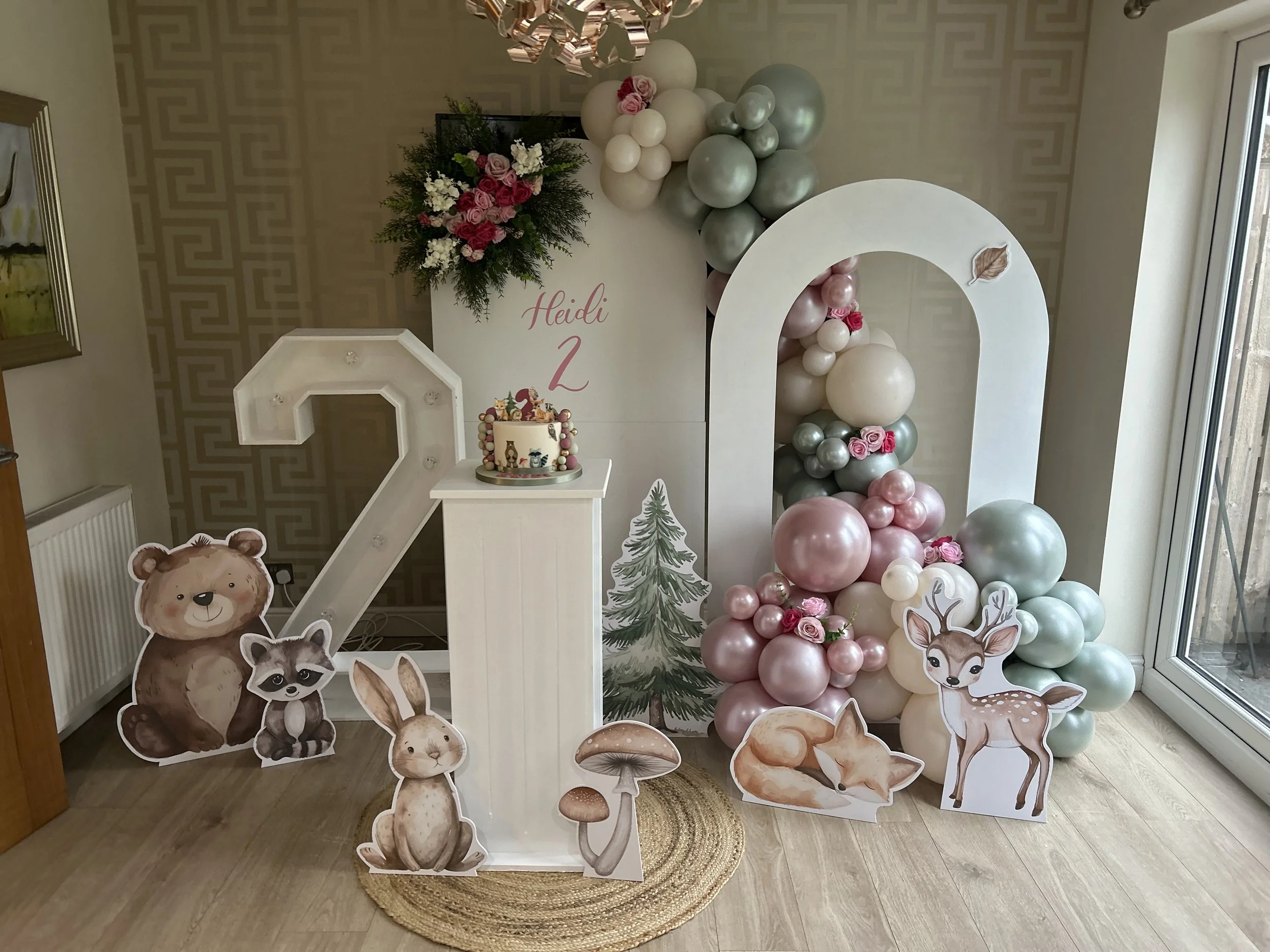 Decorative spring or woodland-themed party area featuring a white arch, pink, white, and green balloons, paper animal cutouts including bear, raccoon, rabbit, deer, and mushroom, a cake with woodland animals, a flower arrangement, and a sign with the