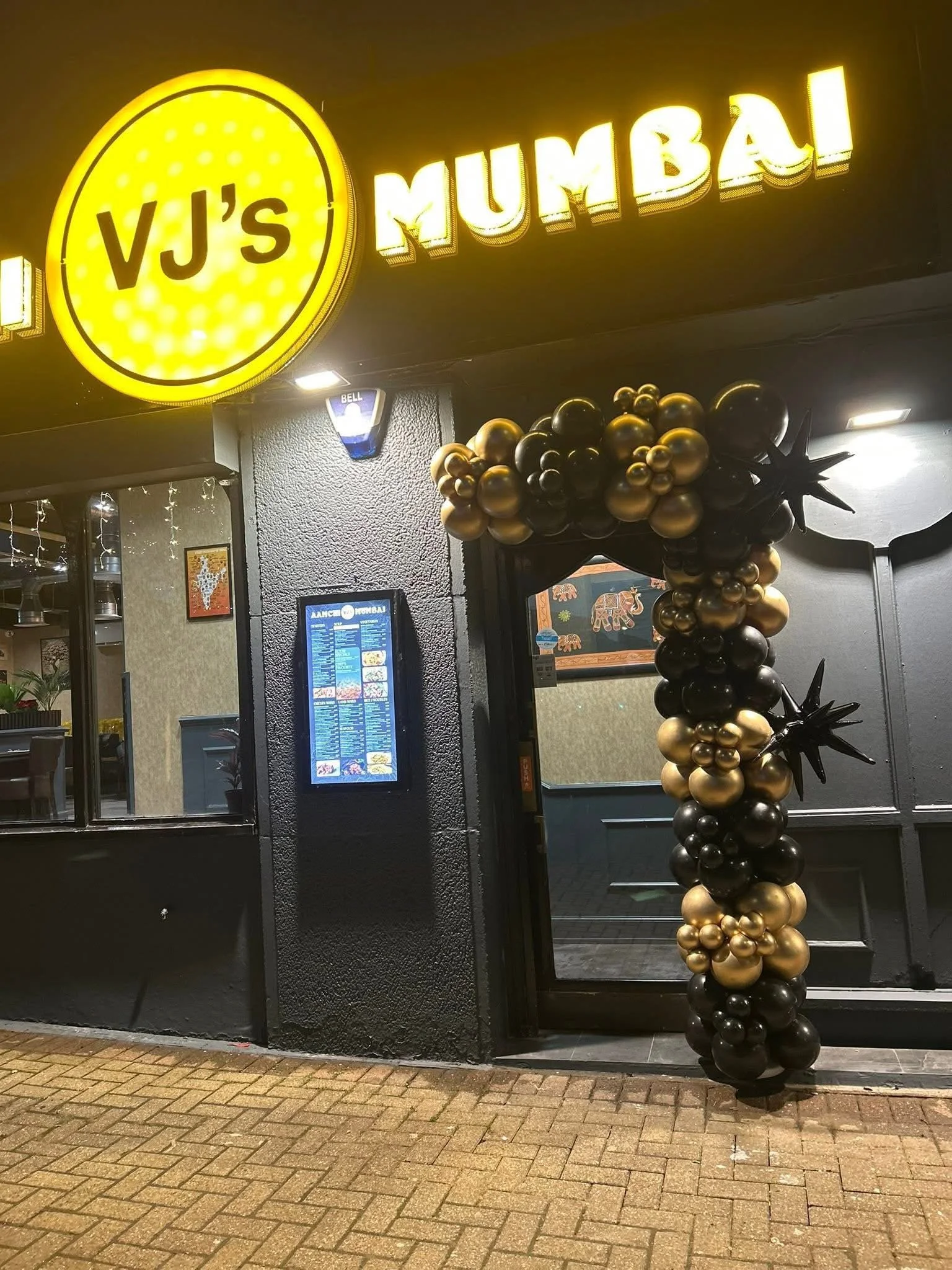 Nighttime exterior of VJ's MUMBAI restaurant, decorated with black and gold balloons and illuminated signage.