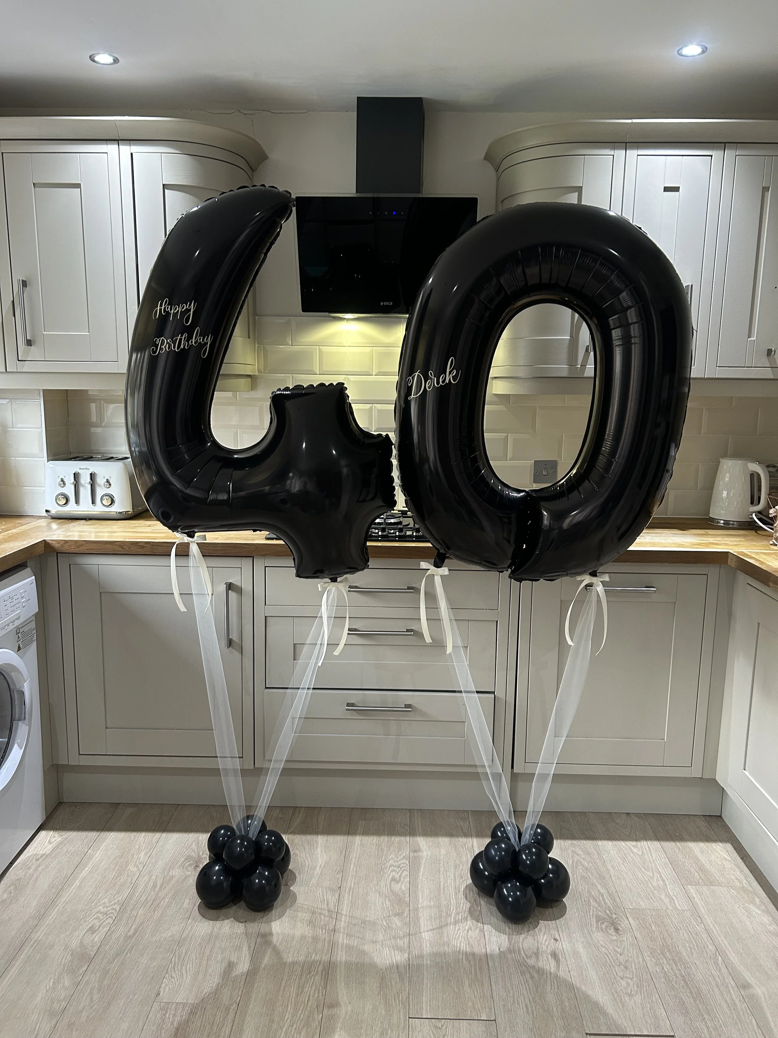 Black balloons forming the number 40 with white writing that says 'Happy Birthday' and 'Derek', balloons anchored with black clusters on either side in a kitchen.
