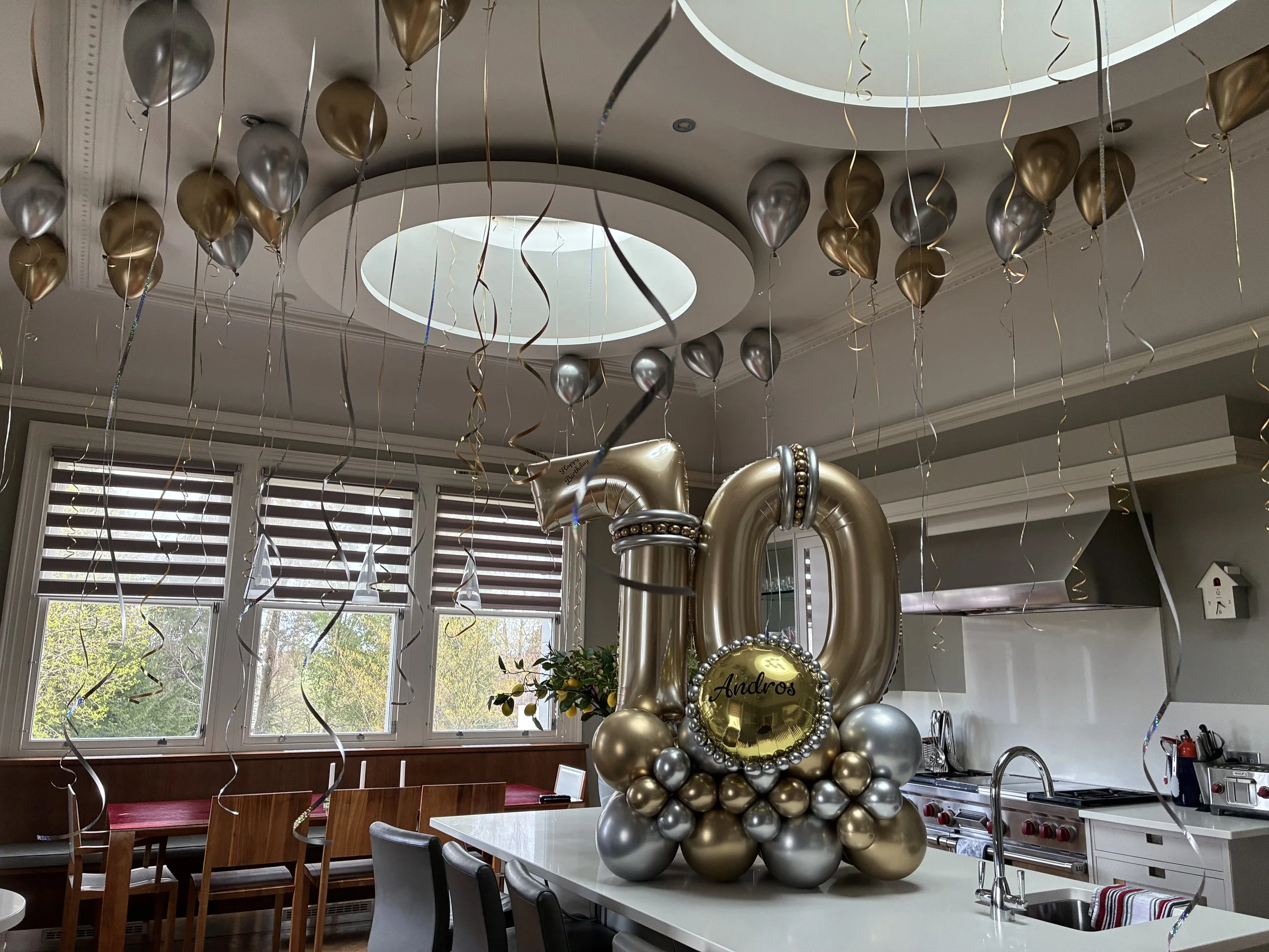 Party decorations inside a bright room with large windows, featuring balloon arrangements in gold and silver, with some balloons spelling out the name 'Andros' amid a grouping of balloons and ribbons hanging from the ceiling.