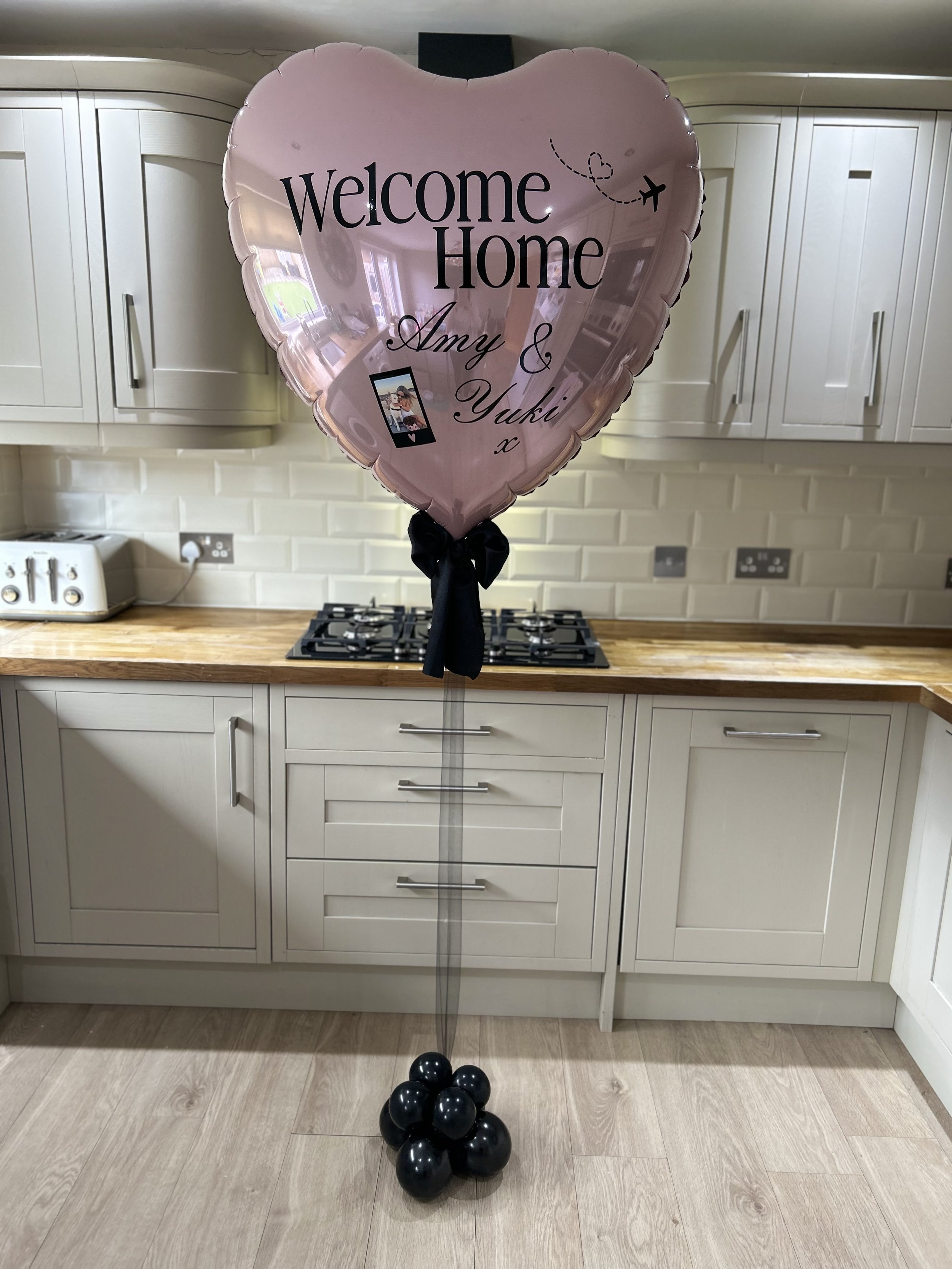 Pink heart-shaped balloon with black text that says "Welcome Home Amy & Yuki" and a small picture of a person taking a selfie, attached to a black ribbon tied at the bottom, set in a kitchen with white cabinets, beige backsplash, and a black stovetop