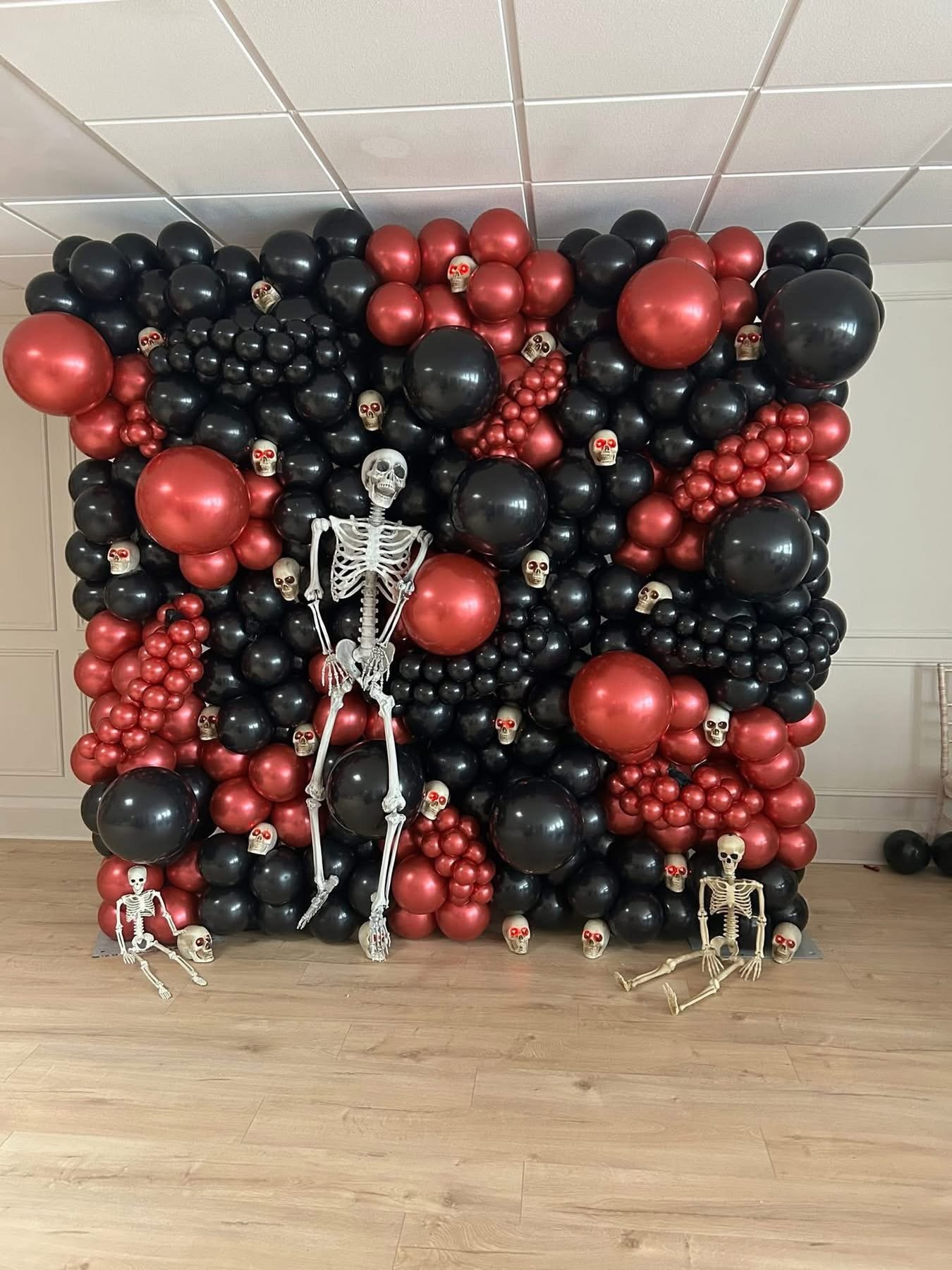 A Halloween decoration wall made of black and red balloons with small skulls attached, and four plastic skeletons posed around the base.