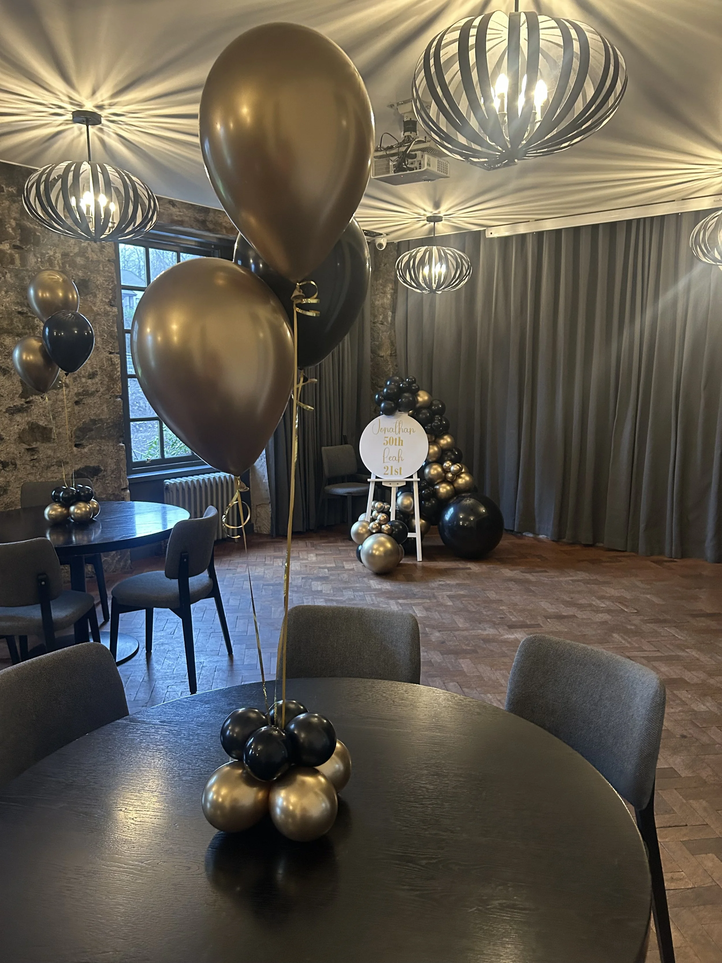 Decorative balloons in black, gold, and silver arranged on a table centerpiece in a celebration setting, with balloons arching and grouped in the background, celebrating a 50th and 21st birthday event.