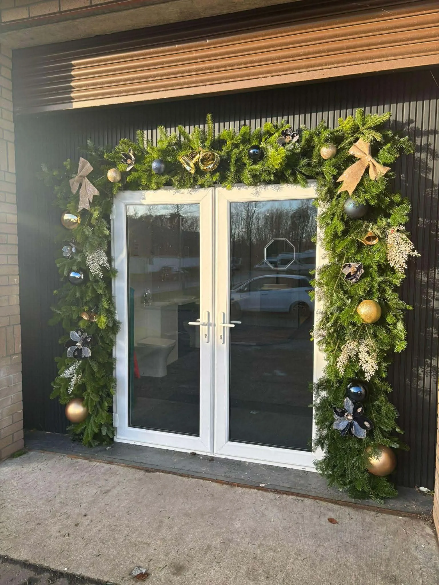 A storefront door decorated with a festive green garland adorned with gold and black ornaments, ribbons, and flowers.