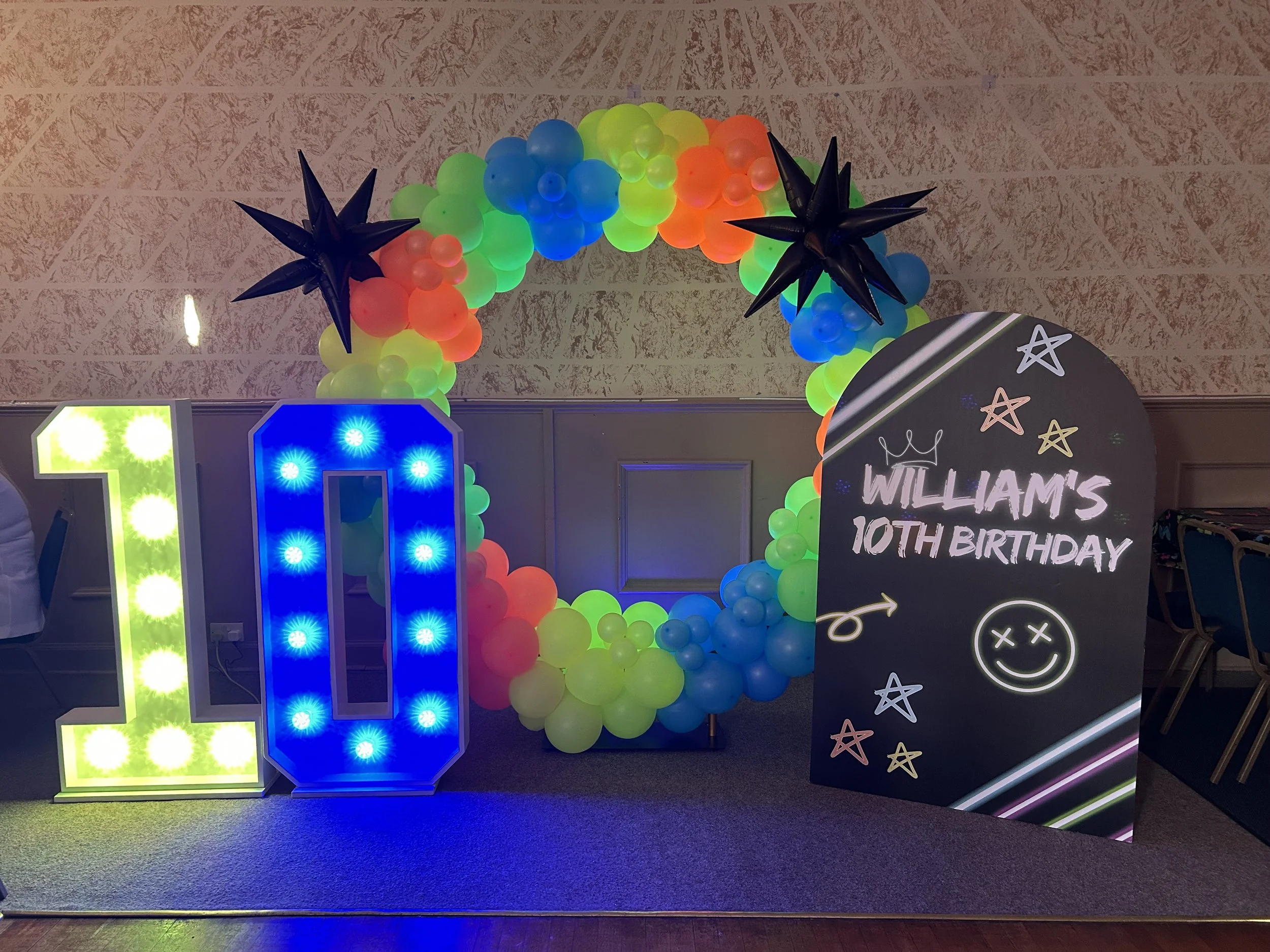 Colorful balloon arch with black star-shaped balloons and green, yellow, orange, blue, and pink balloons at a birthday celebration with a sign reading "William's 10th Birthday" and decorative stars.