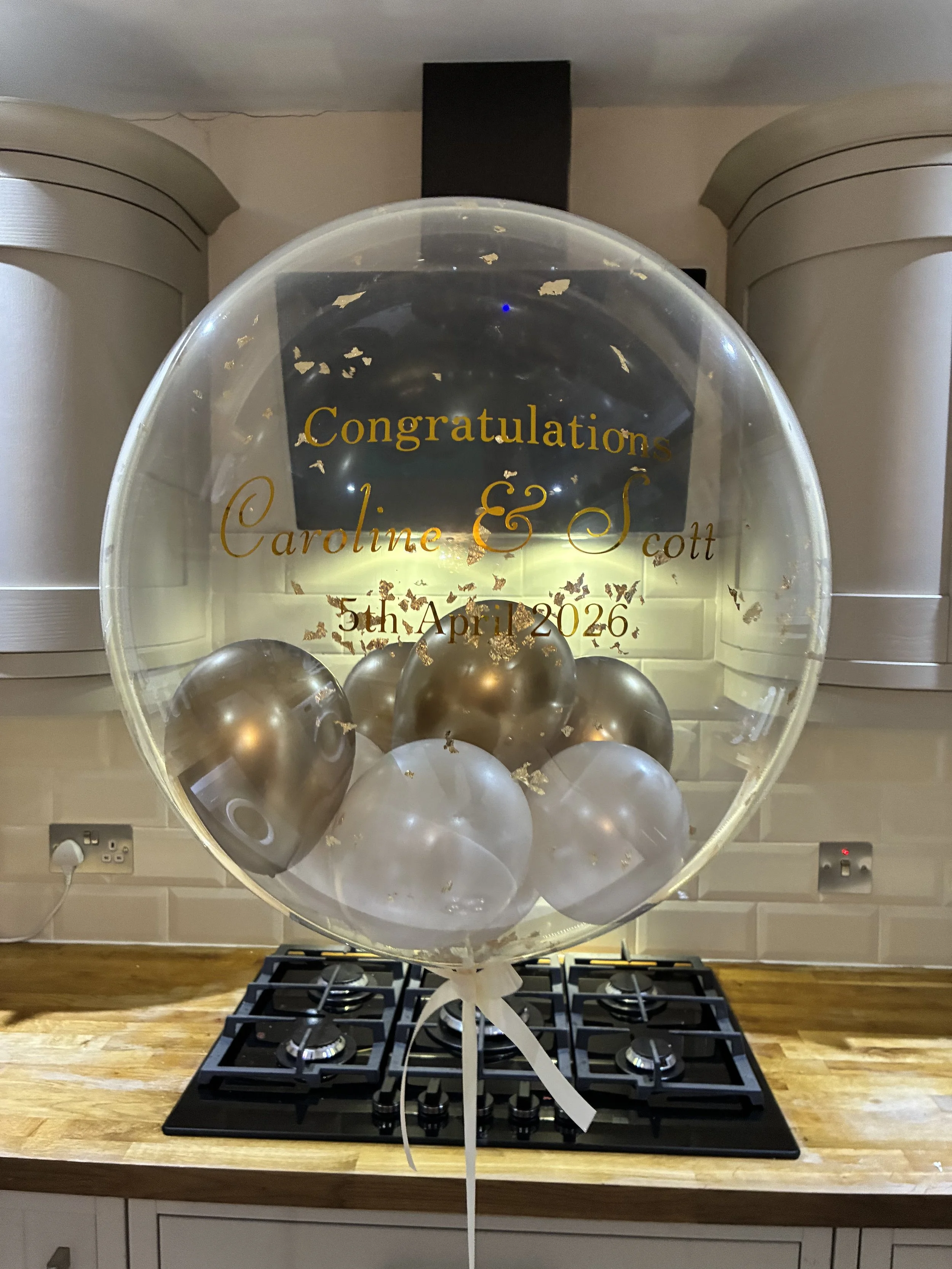 A balloon decoration with gold and silver balloons inside a transparent balloon with gold foil flakes, placed on a kitchen stove with a wooden countertop.