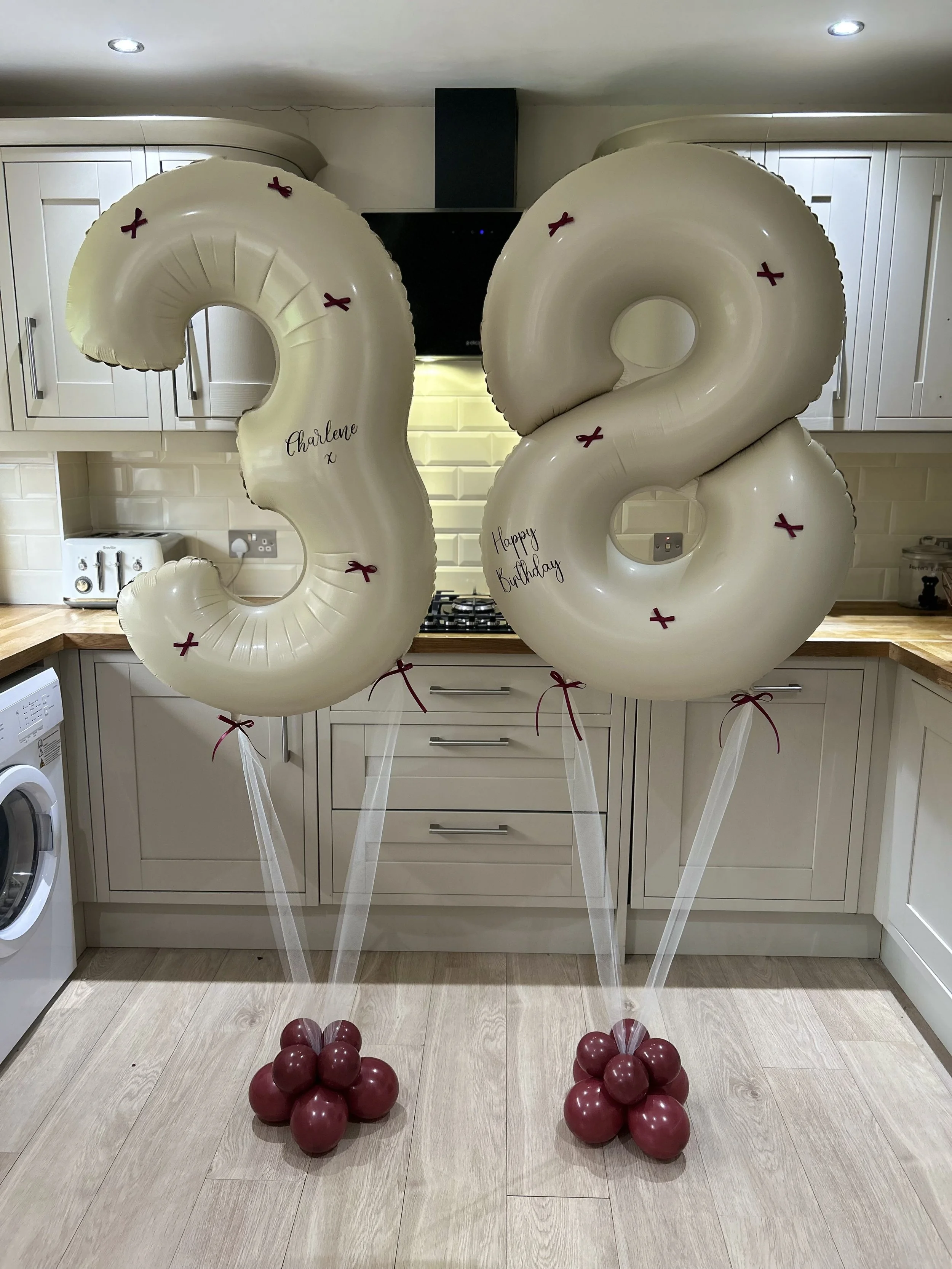 Large white balloons shaped as number 3 and 2 with red ribbons and 'Charlene' and 'Happy Birthday' written on them, surrounded by smaller maroon balloons on a kitchen floor.