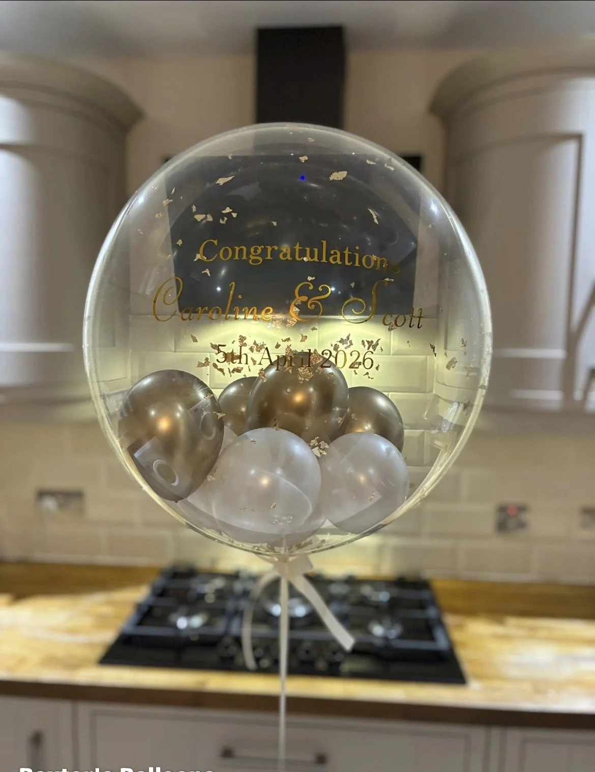 A transparent balloon filled with metallic silver and clear smaller balloons and gold confetti, with a message congratulating Caroline and Scott for their 5th anniversary on April 5th, 2026.
