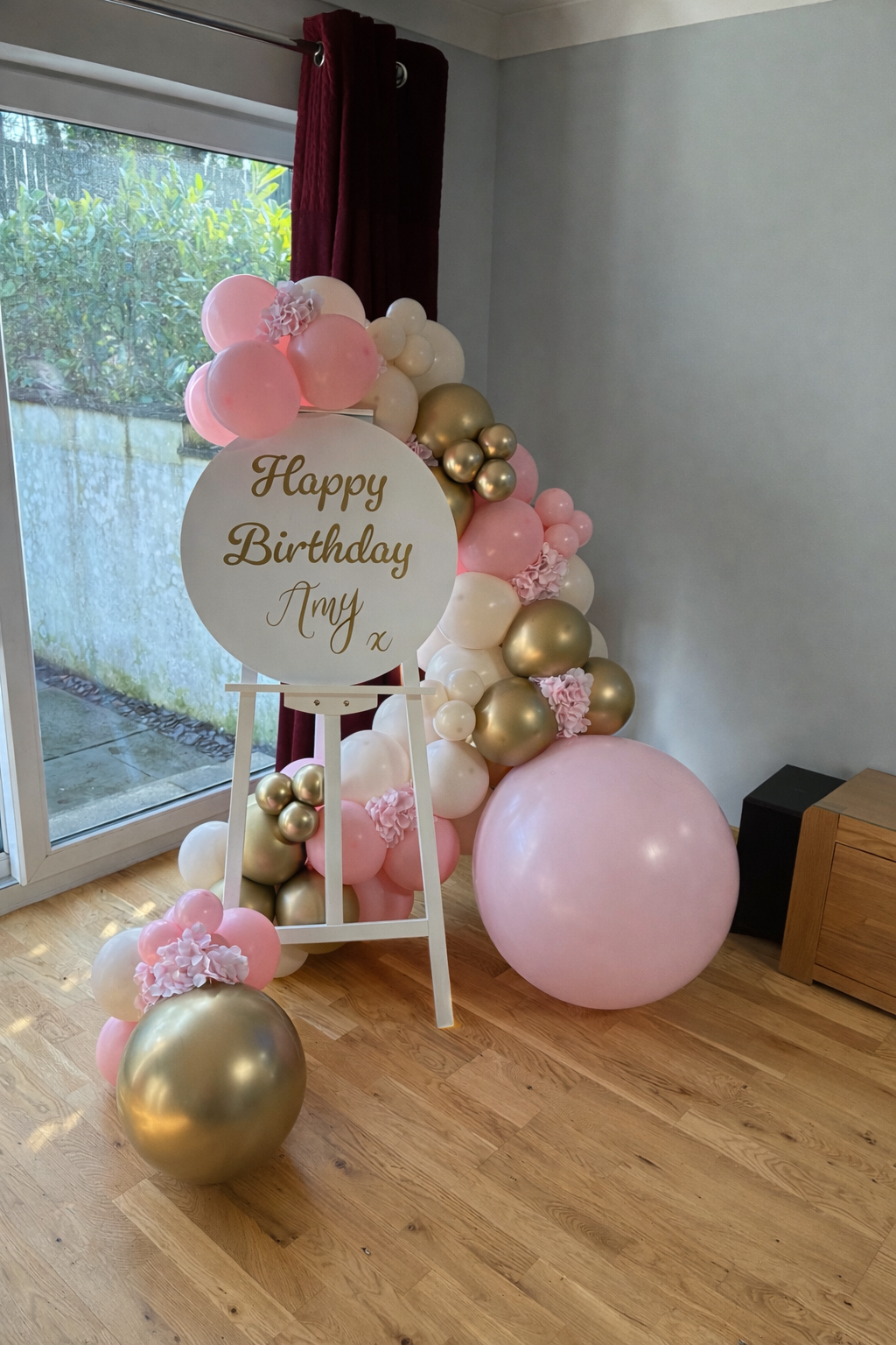 Decorative birthday balloon arrangement with pink, white, gold, and pearl balloons, featuring a sign that reads "Happy Birthday Amy" on an easel by a sliding glass door with curtains in a room with wooden flooring.