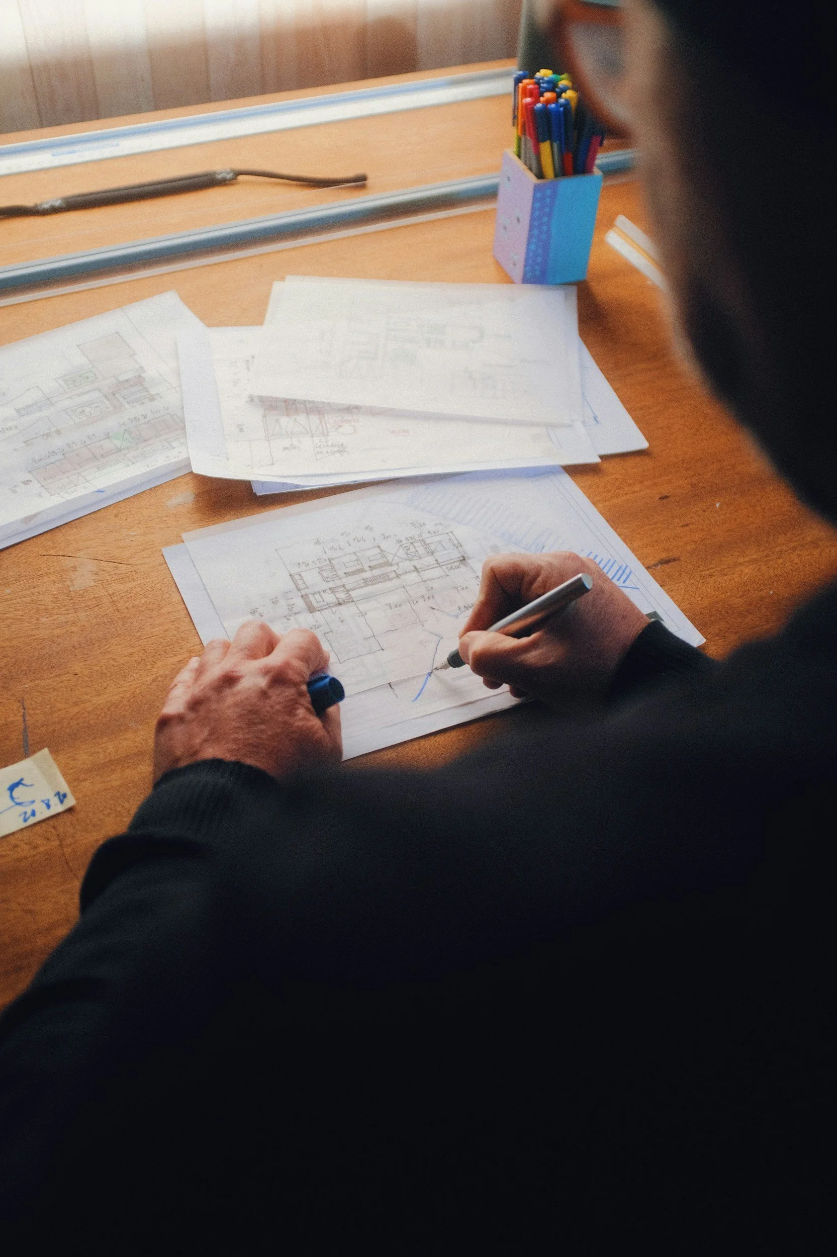 Close-up van een persoon die technische tekeningen en bouwplannen op tafel bekijkt, met potlood en pen in de hand, en een pot met gekleurde stiften op de achtergrond.