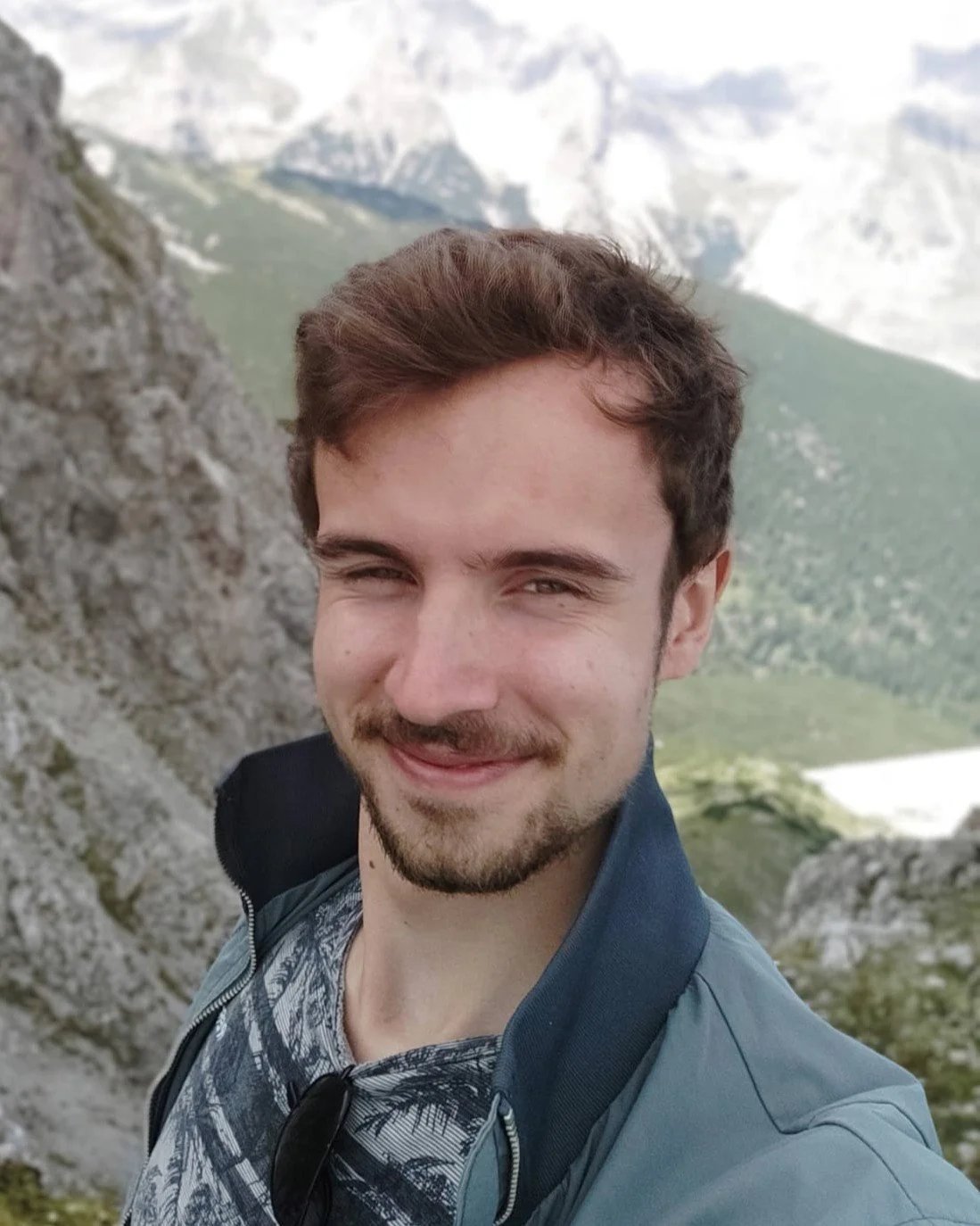 Een jonge man met bruin haar en een lichte baard, die een glimlach vertoont, staat voor een berglandschap met hoge bergen en groene valleien.