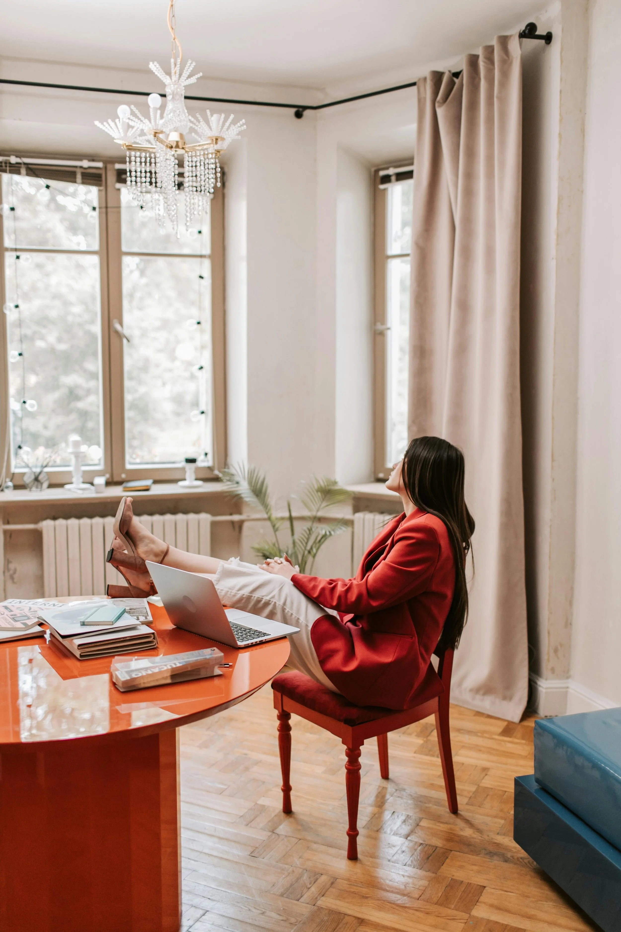 A woman in a red blazer and white pants is sitting on a red chair at a wooden table, with her feet up on the table, working on a laptop. The table has papers, magazines, and a journal on it. The room has large windows with beige curtains, a crystal chandelier hanging from the ceiling, and wood flooring.