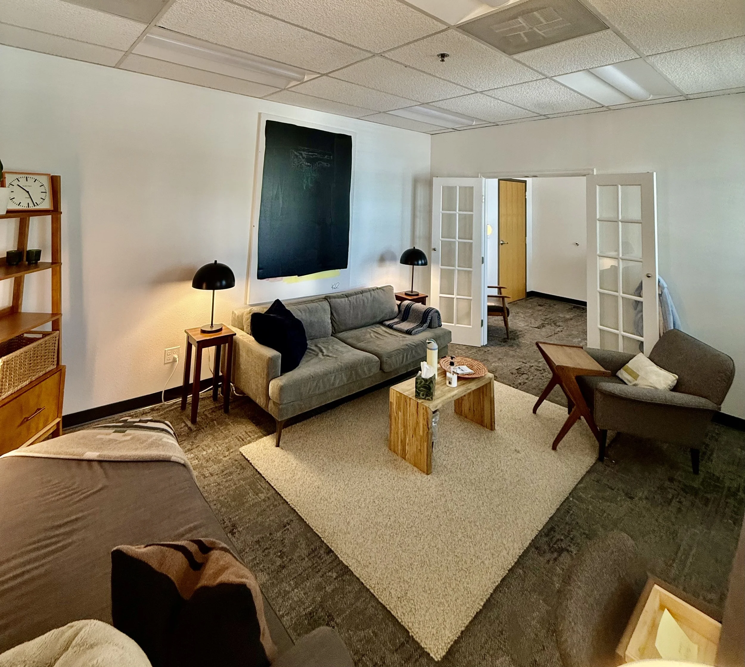Living room with a gray sofa, black lamps, a wooden coffee table, and a carpeted floor. There are white doors with glass panels, a blackboard on the wall, and part of a bed visible in the foreground.