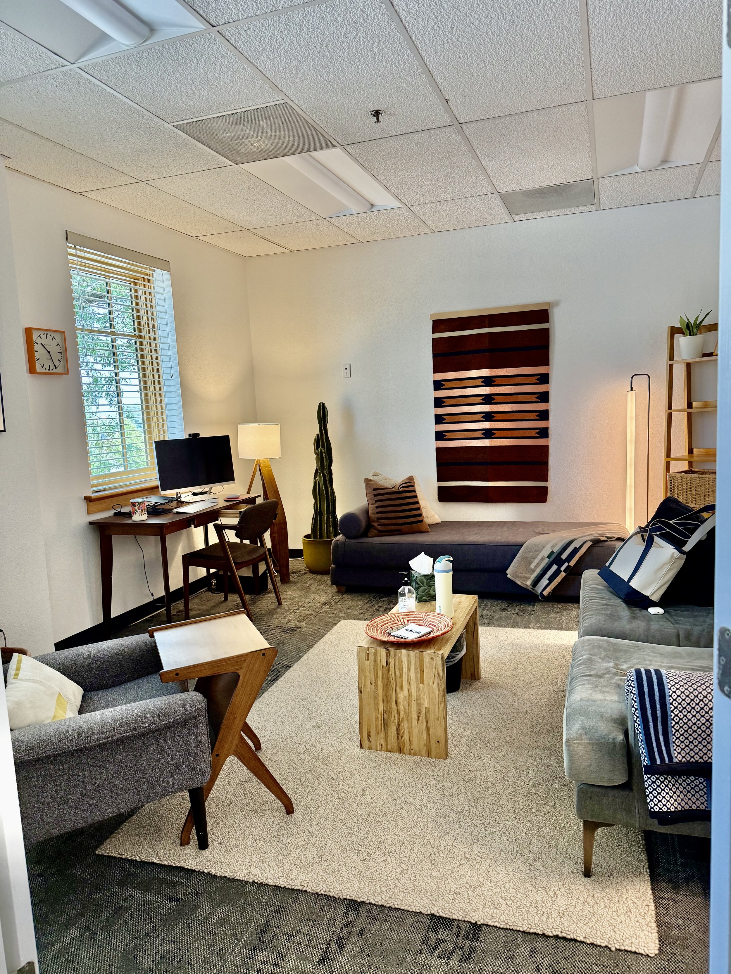 A cozy office or living room space with multiple chairs, a sofa, a small wooden table, a desk with computer, and various decorations including a cactus, a lamp, and wall art. Natural light from a window illuminates the room.