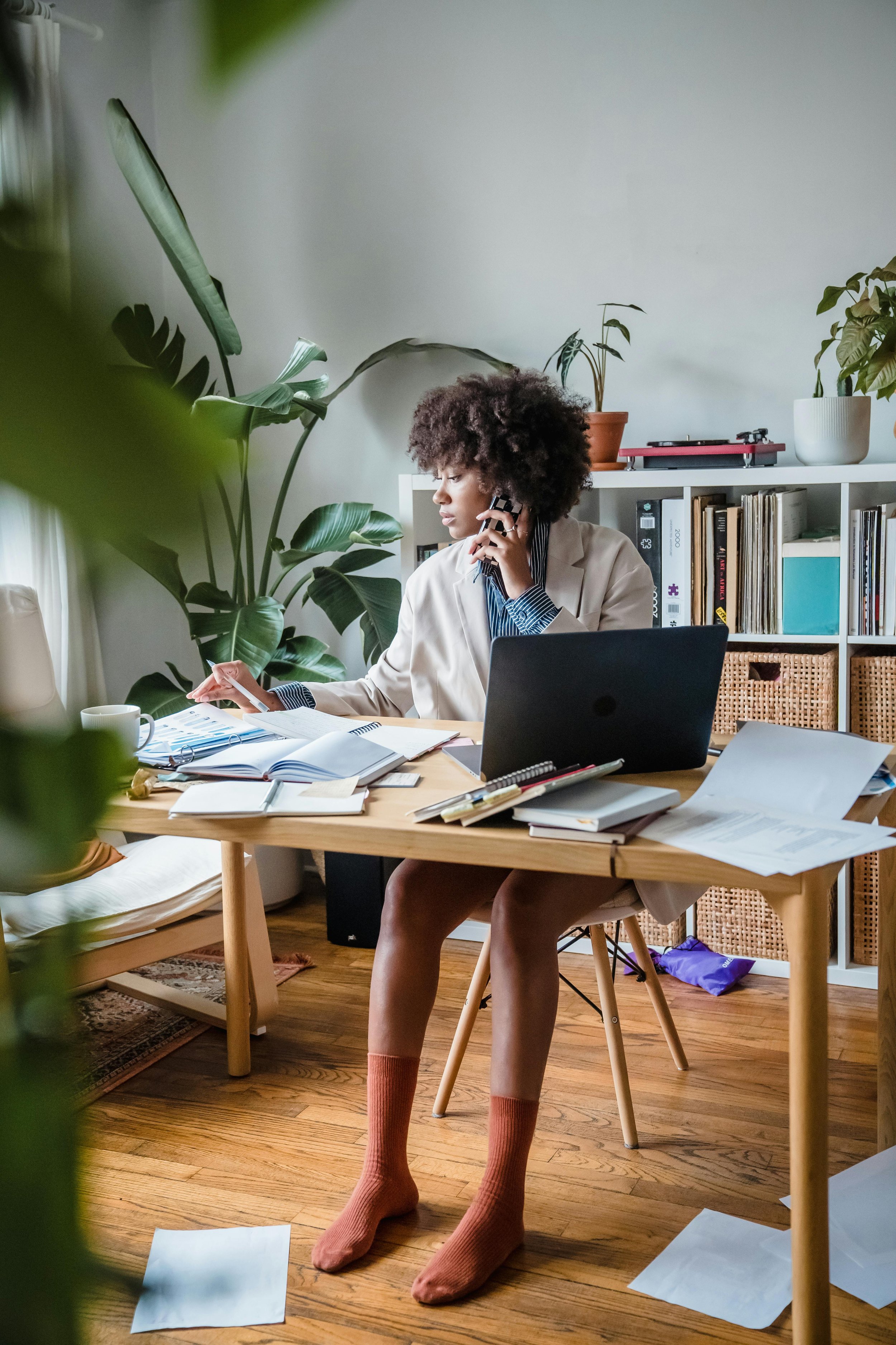 A woman with curly hair wearing a beige blazer, striped shirt, pink socks, and no shoes, sitting at a cluttered wooden desk in a home office, talking on the phone and looking at papers with plants and books on shelves behind her.