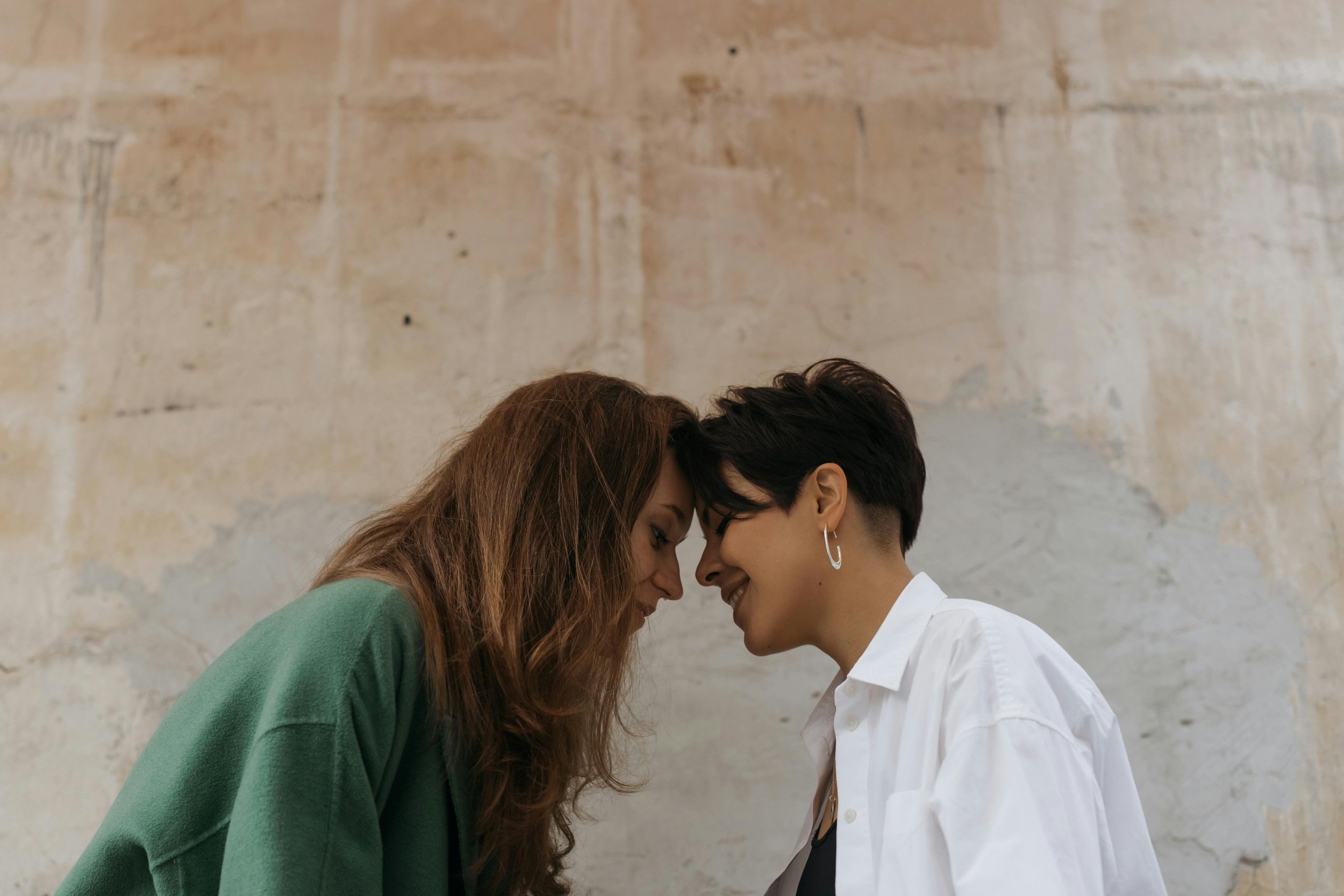 A woman and a woman sharing a moment with their foreheads touching, smiling. The woman on the left has long reddish hair and wears a green sweater. The woman on the right has short dark hair, earrings, and is dressed in a white shirt.