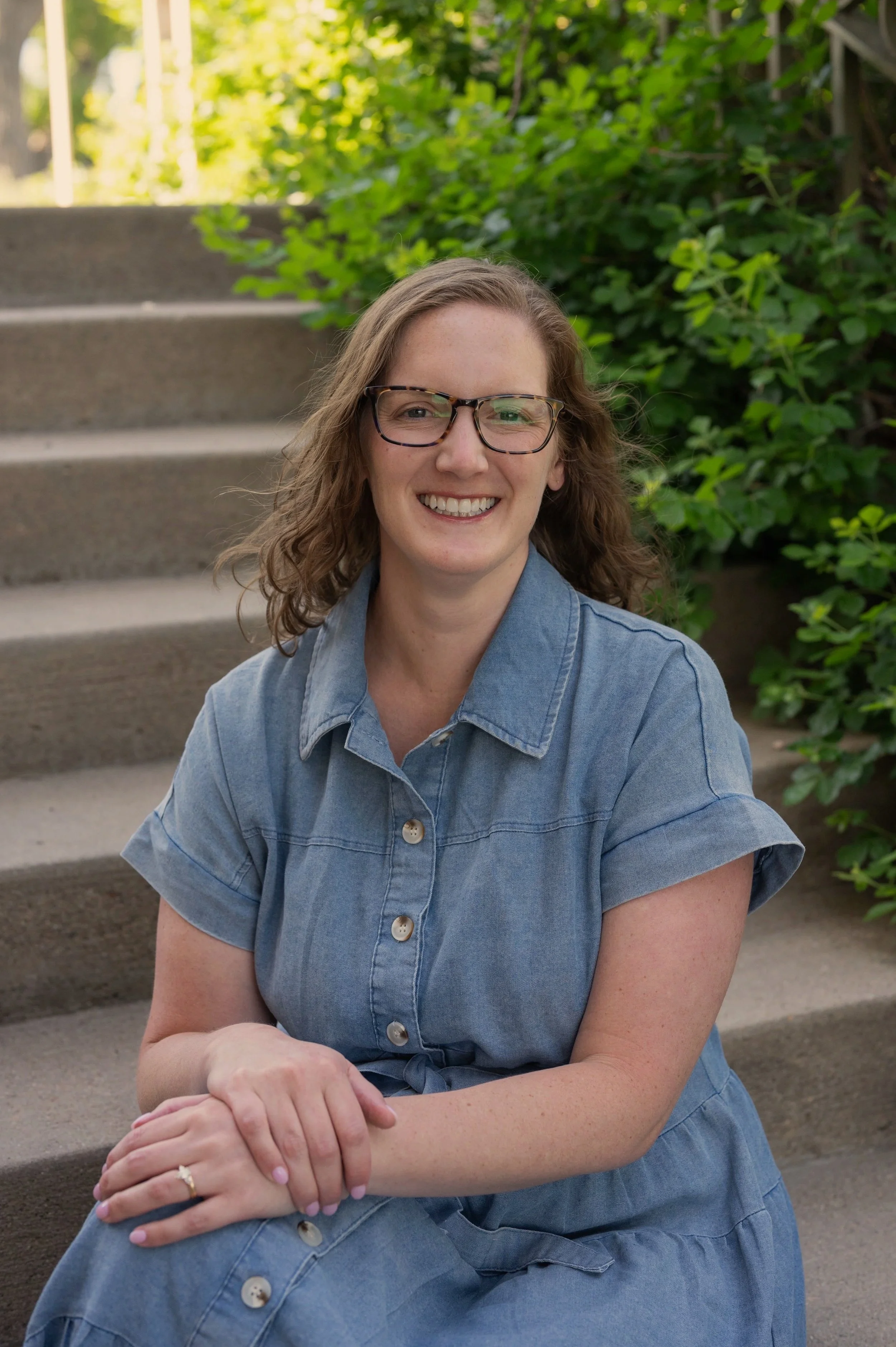 A smiling woman with glasses wearing a denim shirt sitting on concrete steps outdoors, with green foliage in the background.