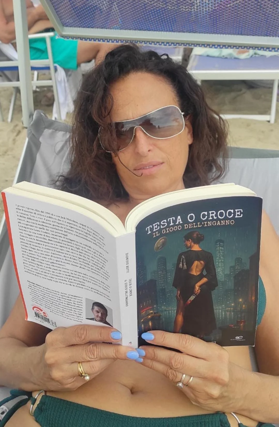 Donna che si rilassa sulla spiaggia leggendo un libro intitolato 'Testa o croce: il gioco dell'inganno'.