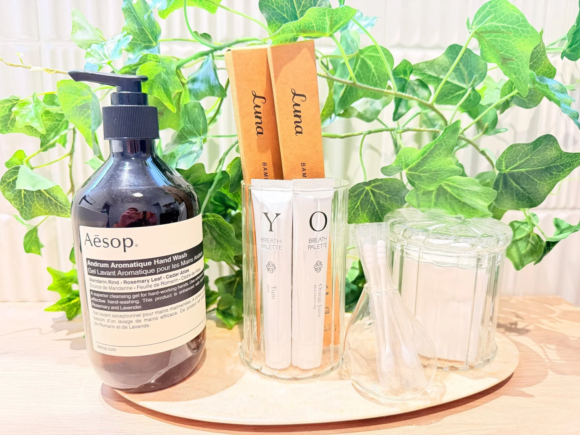 A view of a table with a black liquid bottle, a wooden toothbrush, a clear glass, and a liquid measuring device placed against a background of green ivy.