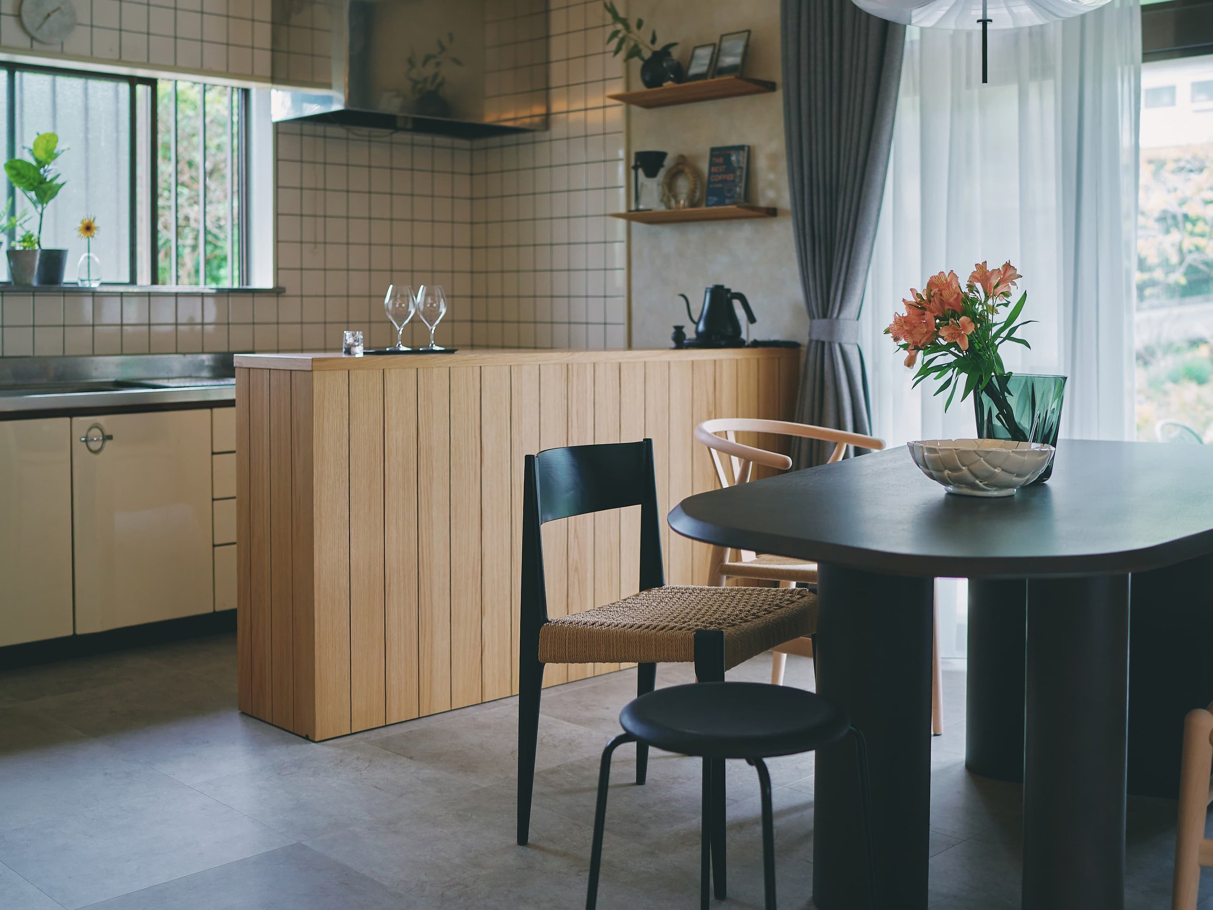 A living room with a wooden counter, behind which are a window and curtains. A vase of pink flowers sits on the table, surrounded by chairs. In the kitchen area, shelves on the wall and cooking utensils such as a coffee maker are visible.