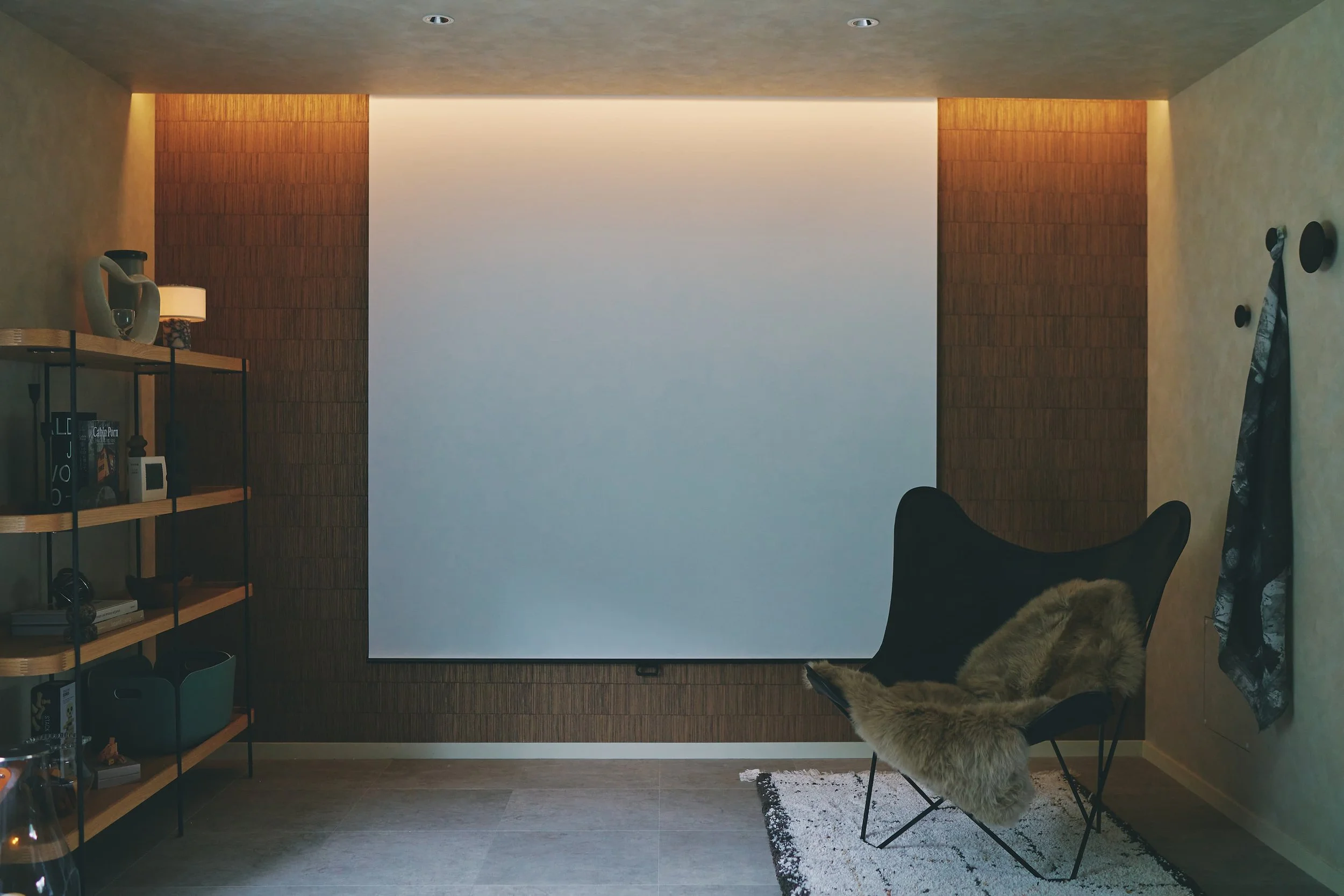 A projector and screen set up on the living room wall. On the left are a bookshelf, some ornaments, and a lamp. On the right are a chair and a fur rug, and hats and bags are hanging on the wall.